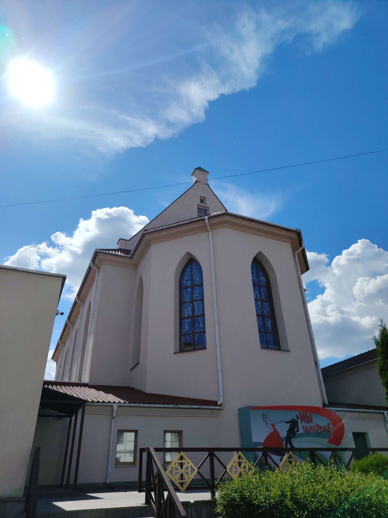 This image presents a vibrant, upward-looking view of a distinctive building, likely a historical structure in Minsk, Belarus, bathed in bright sunlight. The central subject is a tall, cream or light beige building featuring a reddish-brown tiled roof and prominent arched windows, which appear to have blue-tinted or patterned glass. Drainpipes are visible along its facade.
The sky is a brilliant blue, adorned with scattered, fluffy white cumulus clouds. The intense sun, positioned in the upper-left quadrant, creates a strong lens flare and illuminates numerous small, bright specks, possibly dust particles or light reflections, in the atmosphere. This suggests a clear, sunny day, likely around midday.
To the left, a plain, light-colored wall of an adjacent building is partially visible. On the right, a darker roofline and a lush green bush indicate another nearby structure. In the immediate foreground, a decorative wooden railing with yellow and brown geometric cutouts runs horizontally, partially obscuring the bush. Steps lead up to a covered entrance area with dark support pillars.
A prominent and colorful mural or sign is affixed to the lower right side of the main building. It depicts a stylized silhouette of a man and a woman dancing, with the Cyrillic text "МЫ ТАНЦУЕМ!" clearly visible. This translates to "WE DANCE!" The mural adds a contemporary artistic element to the traditional architecture. The scene conveys a bright, pleasant atmosphere in an urban setting.