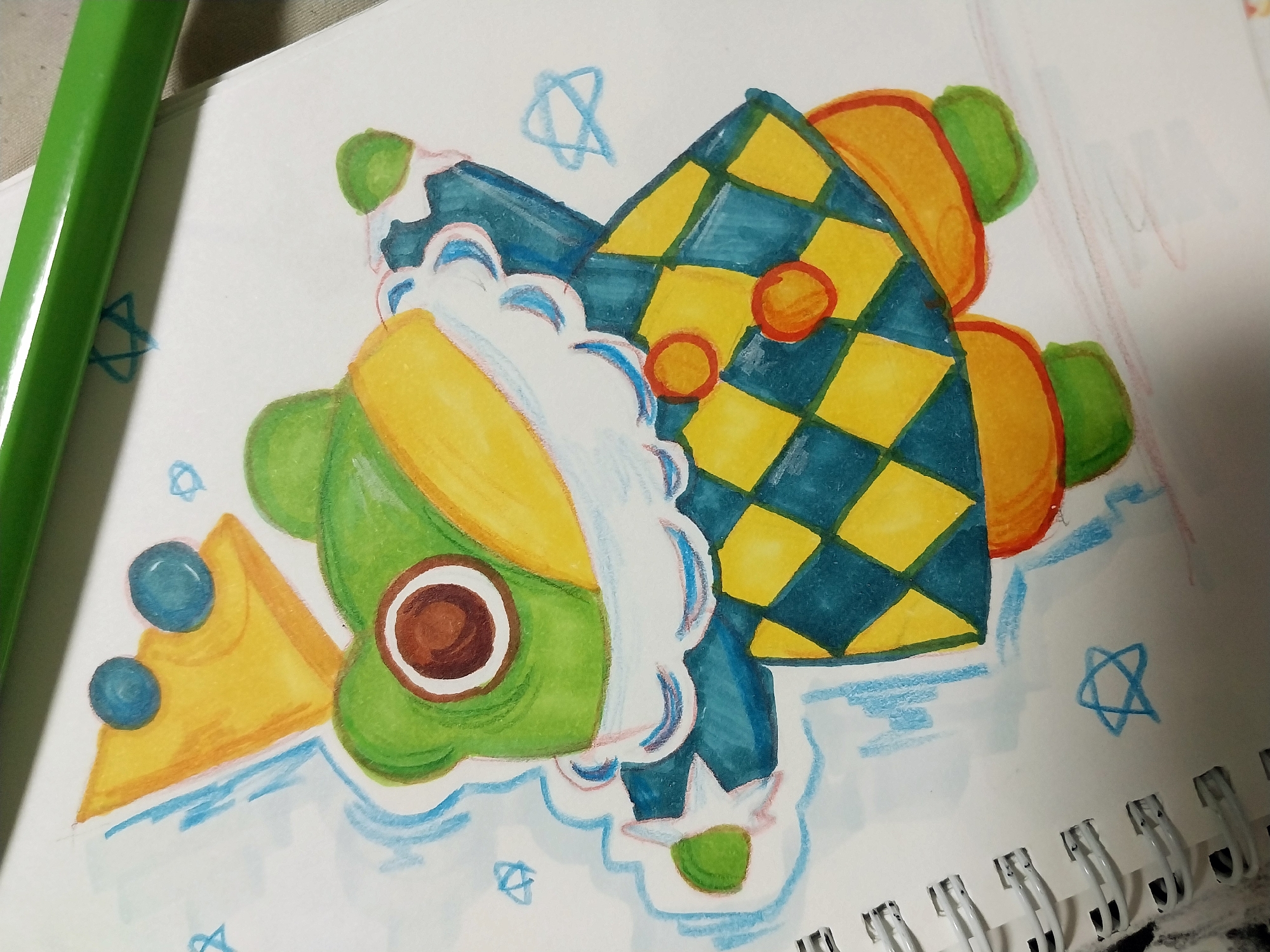 The image is a close-up, overhead shot of a drawing on white paper. The drawing is of a cartoon frog wearing a crown and a checkered outfit. The frog is predominantly green and yellow, with a blue and yellow checkered vest and orange accents. The background is white paper, with some light blue crayon scribbles in the shape of stars and lines. A green marker pen lies horizontally across the top of the image. The drawing appears to be a child's artwork. There is no identifiable location in Gimhae-si, South Korea.