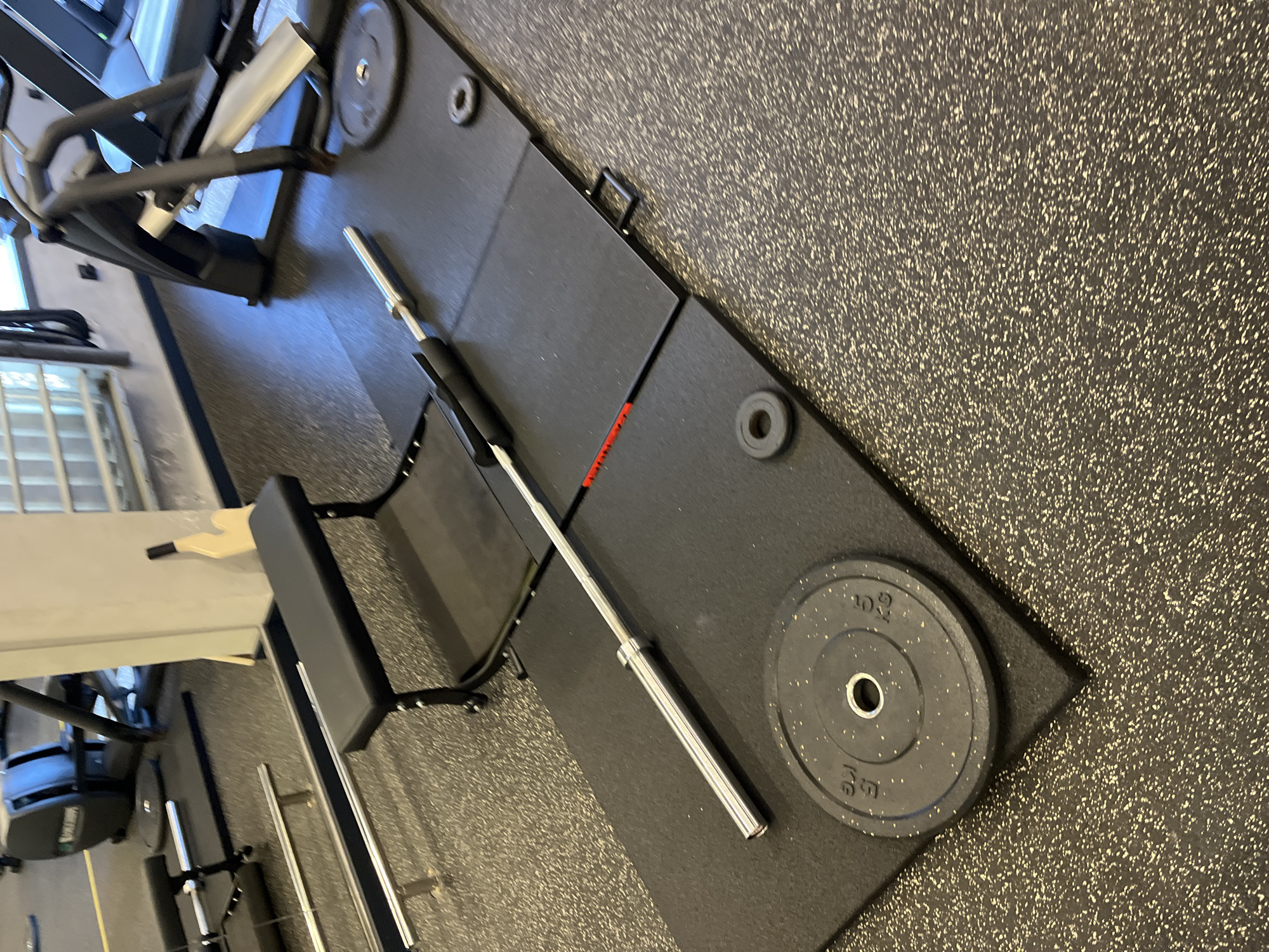 This image captures an interior view of a fitness facility located in Sochi, Russia. The scene primarily focuses on a weightlifting setup on a black rubber mat platform, contrasting with the speckled grey and black rubber gym flooring. A silver Olympic barbell rests horizontally on the platform, with a black 15 KG weight plate positioned on the left side and a black 5 KG weight plate on the right side. Two small black rubber rings are also visible on the mat near the right weight plate. A black workout bench, with a padded seat and backrest, is situated on the mat to the left of the barbell. A small red line marking is visible on one of the mat segments beneath the barbell's midsection, and a black handle is on the edge of another mat segment. In the background, various gym equipment is present, including a rowing machine, a treadmill or elliptical, and a white structural column with a light-colored, possibly wooden, object leaning against it. No individuals are depicted in the scene.