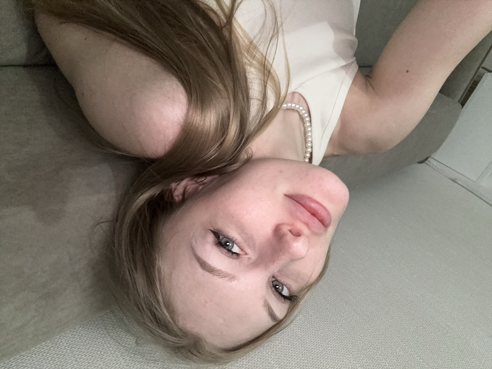 A young Caucasian woman with blonde hair and blue eyes is seen in a close-up, selfie-style shot. She is wearing a white tank top and a pearl necklace. Her head is tilted to her left, and she is looking towards the camera. Her lips are slightly parted, and she appears to be smirking. Her hair is long and wavy, and it falls over her right shoulder. The background is a textured, light gray surface, possibly a ceiling or wall. The lighting is soft and diffused, creating a gentle glow on her face. There are no discernible features of the city of Perm, Russia, visible in the image.