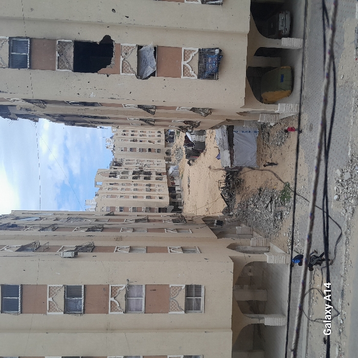 The image depicts a devastated urban landscape in Khan Yunis, Palestinian Territory, characterized by war-damaged residential buildings and a debris-strewn central area. Two prominent, multi-story, light-colored apartment buildings with brown accents and decorative white window frames flank the scene, one on the left and one on the right. Both exhibit significant structural damage, including shattered windows, exposed interiors, and large holes in facades, particularly a substantial impact hole on an upper floor of the building on the right. Air conditioning units protrude or hang damaged from the building exteriors.
The ground between these buildings is a sandy, open expanse heavily littered with rubble, twisted metal, and miscellaneous debris. Several makeshift shelters constructed from tarpaulins and salvaged materials are visible across this area, alongside what appears to be the charred remains of a structure or vehicle. In the midground, three small figures, likely children, are sparsely distributed across the sandy terrain. In the immediate foreground, two darker-clad figures are positioned on a paved, debris-scattered road, partially obscured by a network of overhead black electrical cables. One figure is bent over. Under the arched ground-floor sections of the right building, a green tarp-covered vehicle and another lighter-colored vehicle are parked. The sky above is overcast with patches of blue.