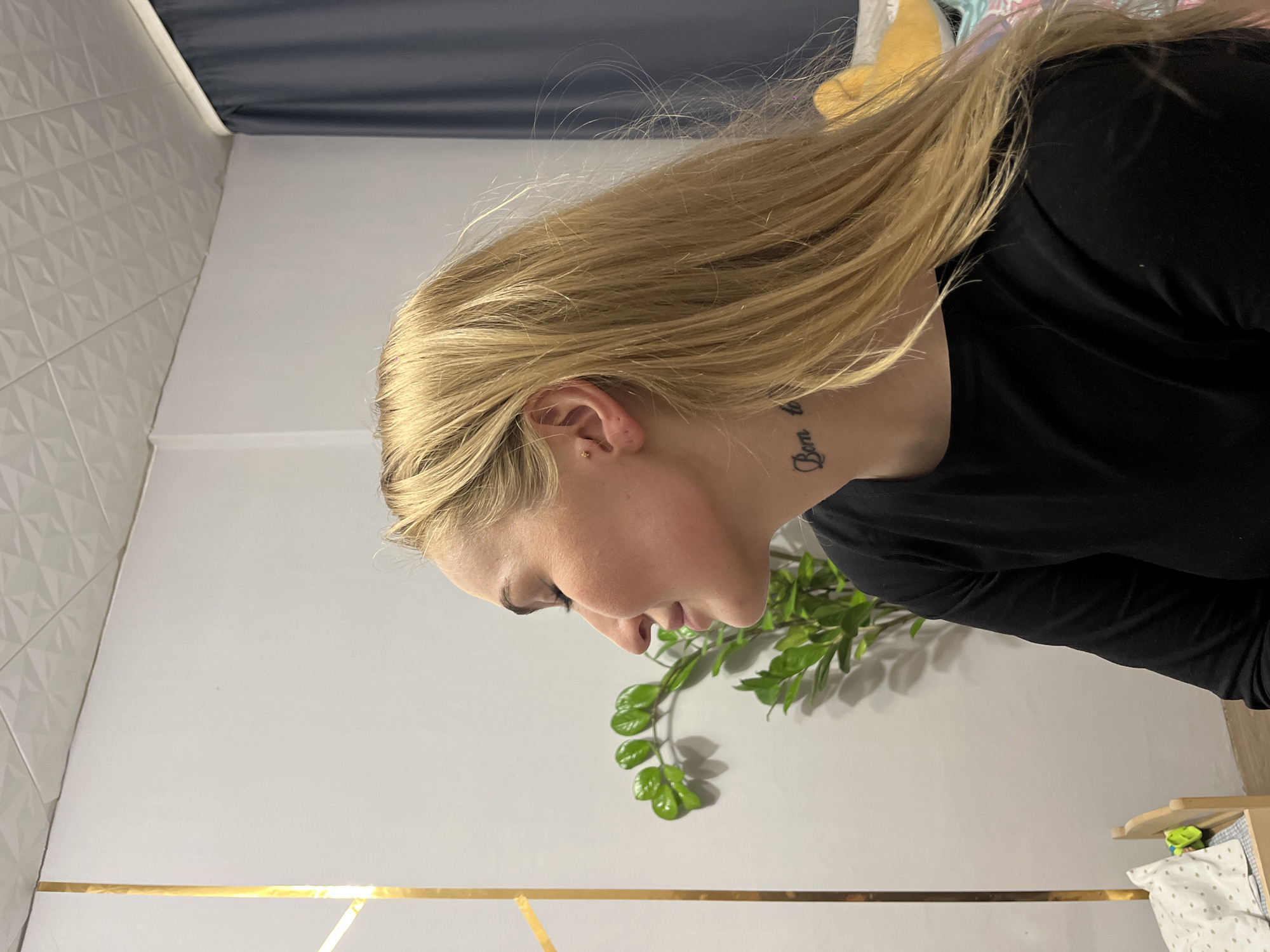 The image displays a head-and-shoulders profile of a blonde female, facing left with her eyes closed or downcast, against an indoor residential background located in Chaikovsky, Russia. The female exhibits fair skin, long blonde hair, and a small gold stud piercing in her right ear. A black cursive tattoo, reading "Born to" followed by partial text, is visible on the right side of her neck, below the ear. She is wearing a plain black long-sleeved garment.
The background consists of a plain white wall on the left, featuring a vertical decorative element of two golden strips forming an acute angle. A green leafy plant is situated behind the subject's head. To the far right, a dark blue or gray curtain hangs. The ceiling is white with a repeating geometric relief pattern. In the lower left foreground, a miniature wooden bed frame is visible, containing a white pillow with brown polka dots and a blue and white checkered blanket. A small green object rests on the miniature bed. The floor visible in the lower left corner appears to be light-colored wood.