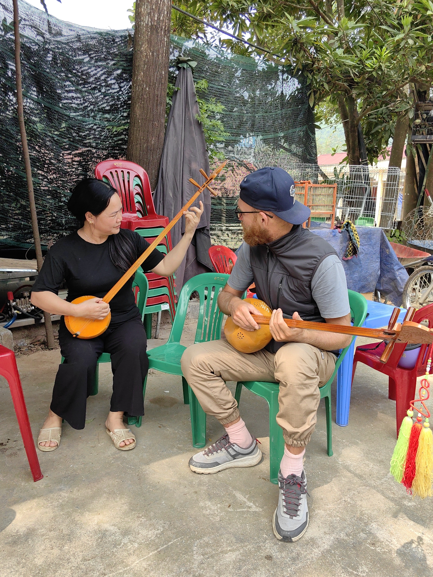 In Niêm Sơn Commune, Vietnam, a woman and a man are seated on plastic chairs, each holding and seemingly playing a T’rưng instrument. The woman, on the left, wears a black shirt and pants with light-colored sandals; she holds her instrument upright, facing the man. The man, on the right, has a beard and wears a baseball cap, gray vest, beige pants, and sneakers with pink socks. He also holds a T’rưng. They appear to be interacting, possibly sharing musical knowledge.
The environment includes a combination of outdoor and covered spaces. Red and green plastic chairs are scattered around. In the background is a black netting-type screen. Behind the man, there is a blue tarp-covered surface and what appears to be a birdcage. A tree is visible in the middle of the scene. The ground is concrete.
