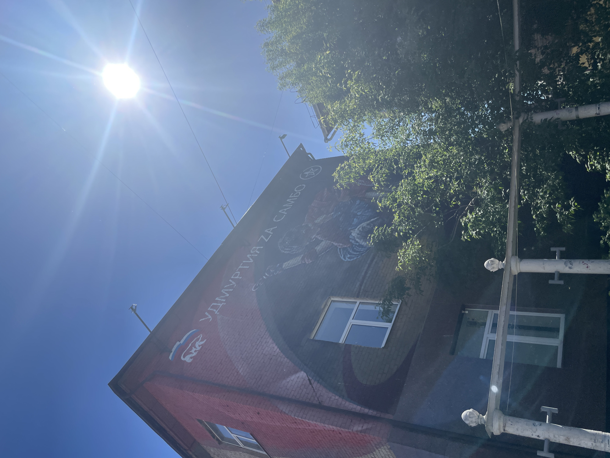 A low-angle shot captures the side of a brick building under a bright, sunny sky in Izhevsk, Russia. A mural adorns the building, with text in Russian reading "Удмуртия за Самбо" (Udmurtia for Sambo). The mural also features a figure in the midst of what appears to be a sambo wrestling move. Windows are visible on the side of the building. A green tree is present, partially obscuring the building. A white metal street fixture with decorative features is in the right foreground.