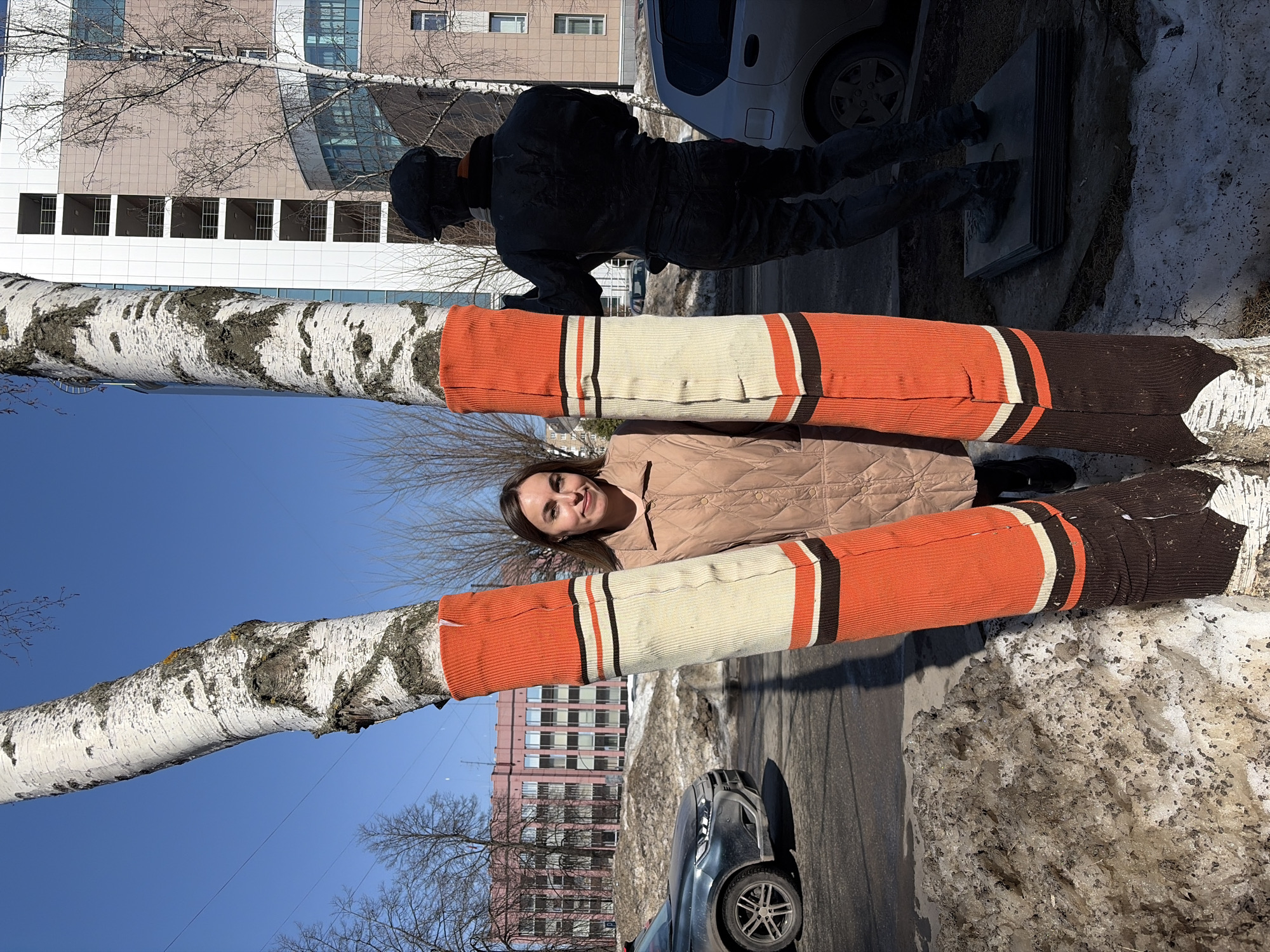 The image depicts a scene in Izhevsk, Russia, likely in a public space near a street with vehicles and buildings visible. A fair-skinned woman with dark hair and a light brown jacket lies between two birch trees. The trees are wrapped in colorful knitted sleeves, featuring orange, beige, and brown stripes. Behind the trees, there's a statue of a figure in dark clothing. The ground shows remnants of melting snow. Buildings with light-colored facades and windows are visible in the background. A car is parked on the street. The sky is blue.