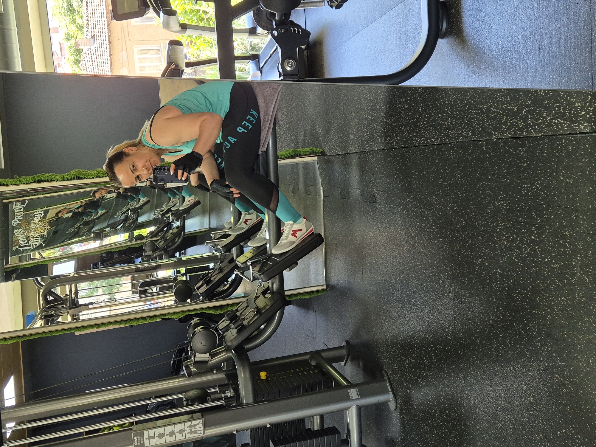 A woman is seated on a weight machine in a gym, taking a selfie with her smartphone. She is wearing a teal tank top, black leggings with teal accents, black fingerless gloves, and white athletic shoes with red and grey details. The gym has a dark grey floor, large mirrors, and various weight machines. A large window in the background reveals an outdoor scene with buildings and trees. The woman is holding a dark-colored bottle in her right hand.