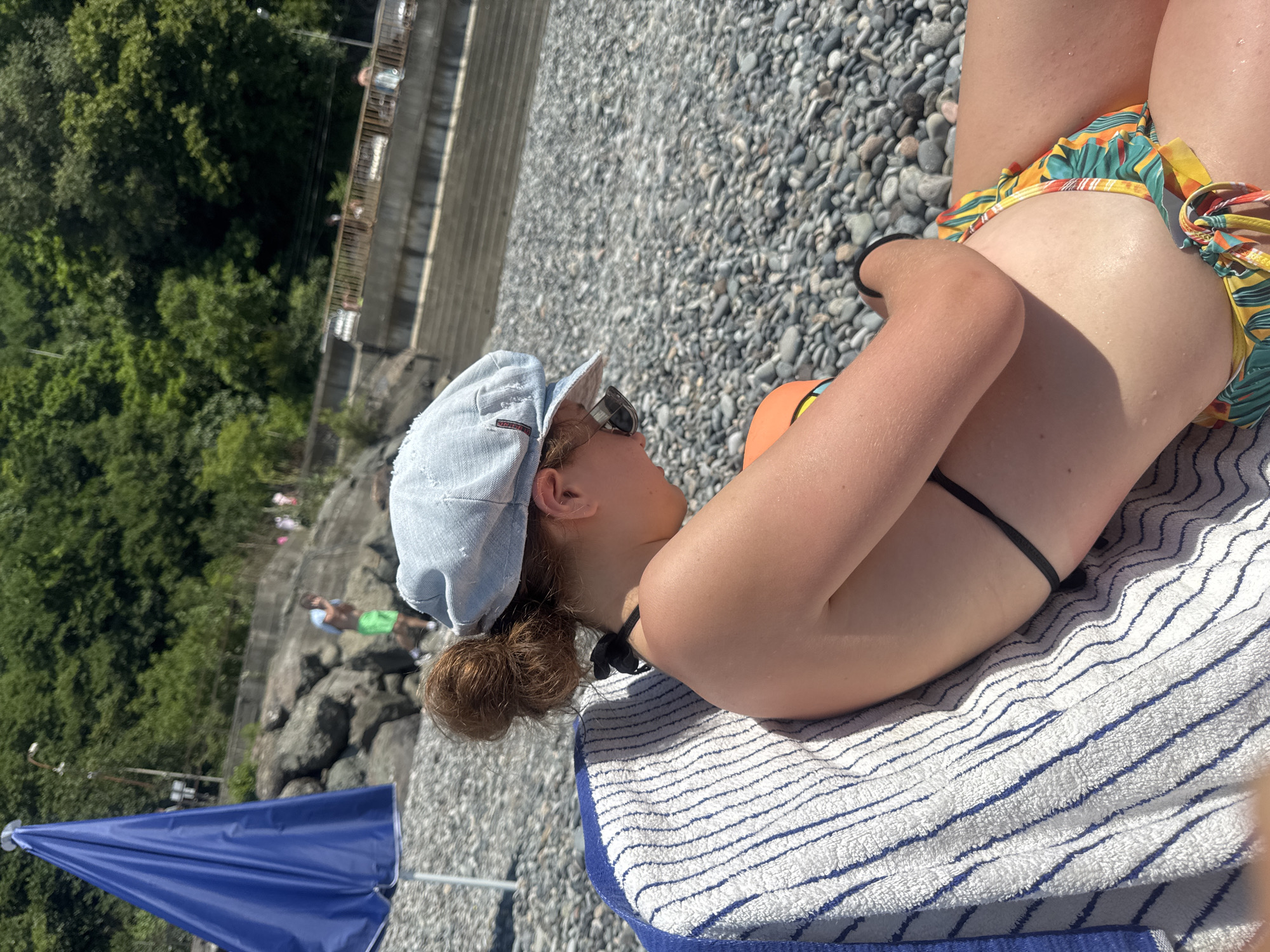 A person lies on a towel on a rocky beach, facing away from the camera and looking towards the water. They are wearing a light blue denim cap, sunglasses, and a colorful bikini with a tropical print. The person's hair is pulled back into a bun. A blue umbrella is visible in the foreground, and in the background, there are stairs leading down to the water, trees, and other people on the beach. The sun appears to be shining, suggesting it is daytime and likely warm weather. The scene evokes a relaxed, vacation-like atmosphere.