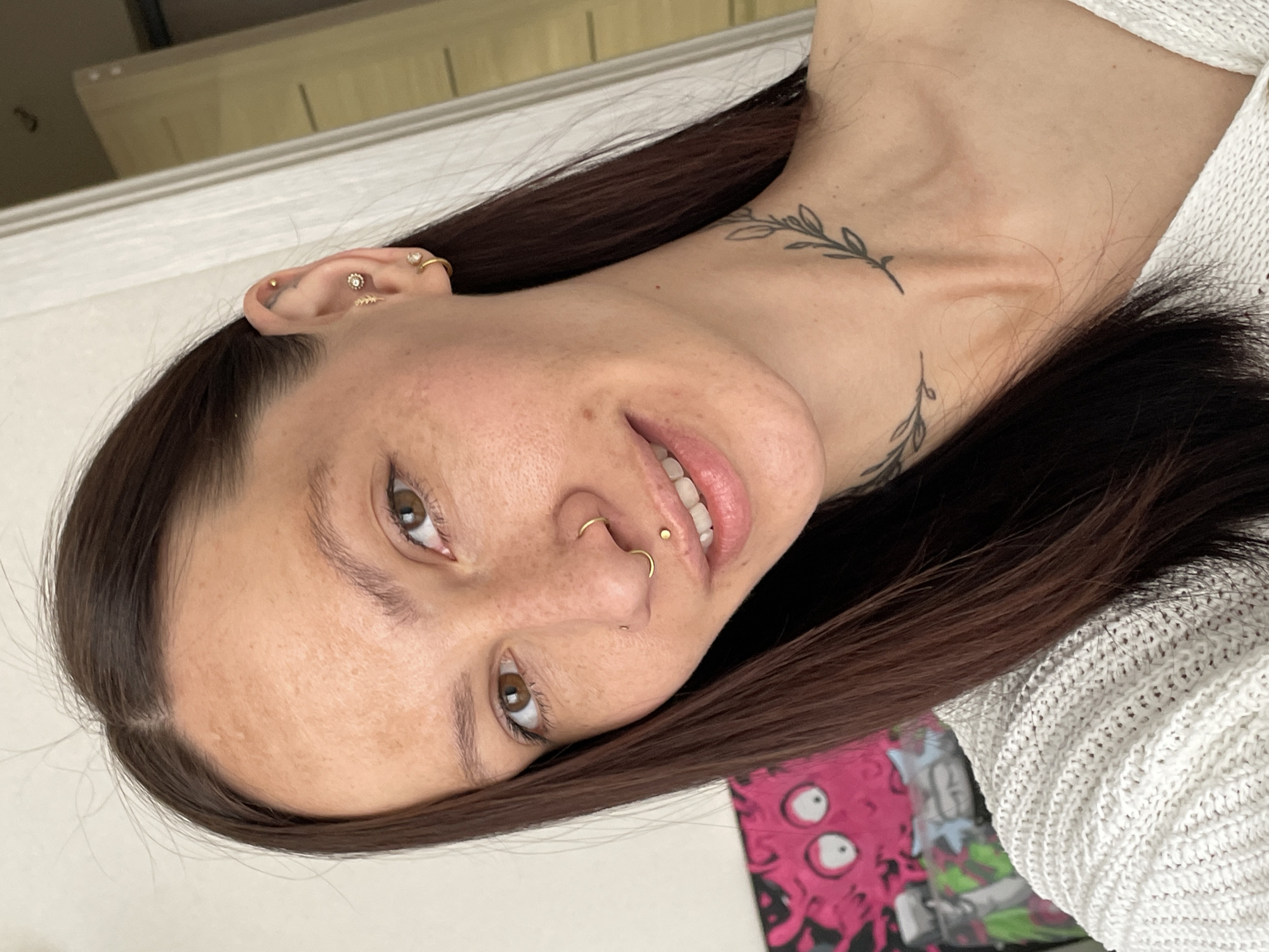 A woman with dark hair is seen from the chest up, angled towards the camera. She has light skin, brown eyes, and multiple piercings in her ear, nose, and lip. A leaf-like tattoo is visible on her neck. She is wearing a white knitted sweater. A poster with cartoonish figures is partially visible in the background. The location is not discernible from the image; the city of Pervomaysky, Russia, cannot be confirmed.
