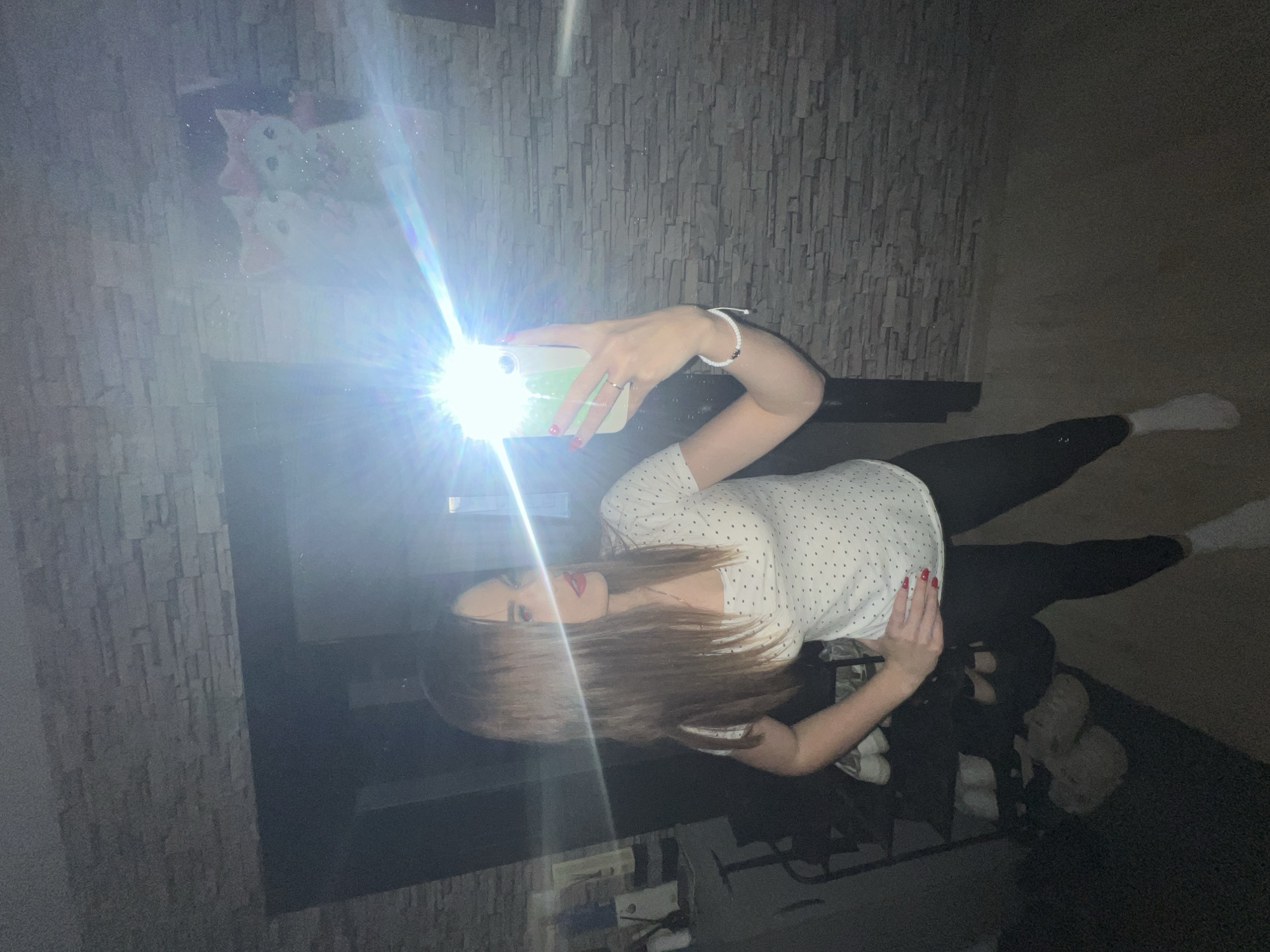 A young woman is taking a selfie in a room. She is holding her phone with her right hand, the flash reflecting off a mirror in front of her. She is wearing a white, polka-dot shirt, and black leggings. She has long, brown hair and red nail polish. The background includes a grey, textured wall and a dark floor. The room appears dimly lit. There is no mention of Noviy, Russia.