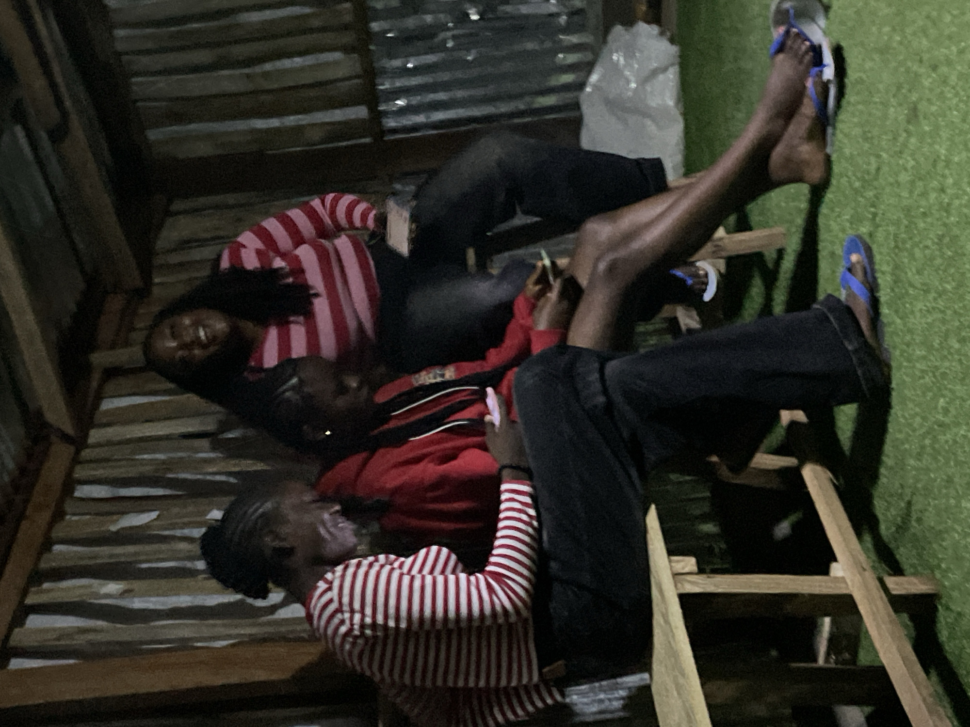 The image depicts an outdoor scene in Durumi, Nigeria, where three young women are gathered. They are seated on wooden benches in what appears to be an open-air shelter with a corrugated metal roof and wooden-slatted walls. The ground is covered in artificial turf.
The woman on the left, wearing a red and white striped long-sleeved shirt and dark pants, is looking down at her phone. Beside her, a woman in a red hooded jacket and dark pants is also looking at her phone. The third woman, positioned at the back and to the right, is wearing a pink and red striped long-sleeved shirt and dark pants. She is looking upwards and laughing. All three women appear to be engaged with their phones or interacting with each other in a relaxed setting. The lighting suggests it is dusk or nighttime, with artificial light illuminating the scene.