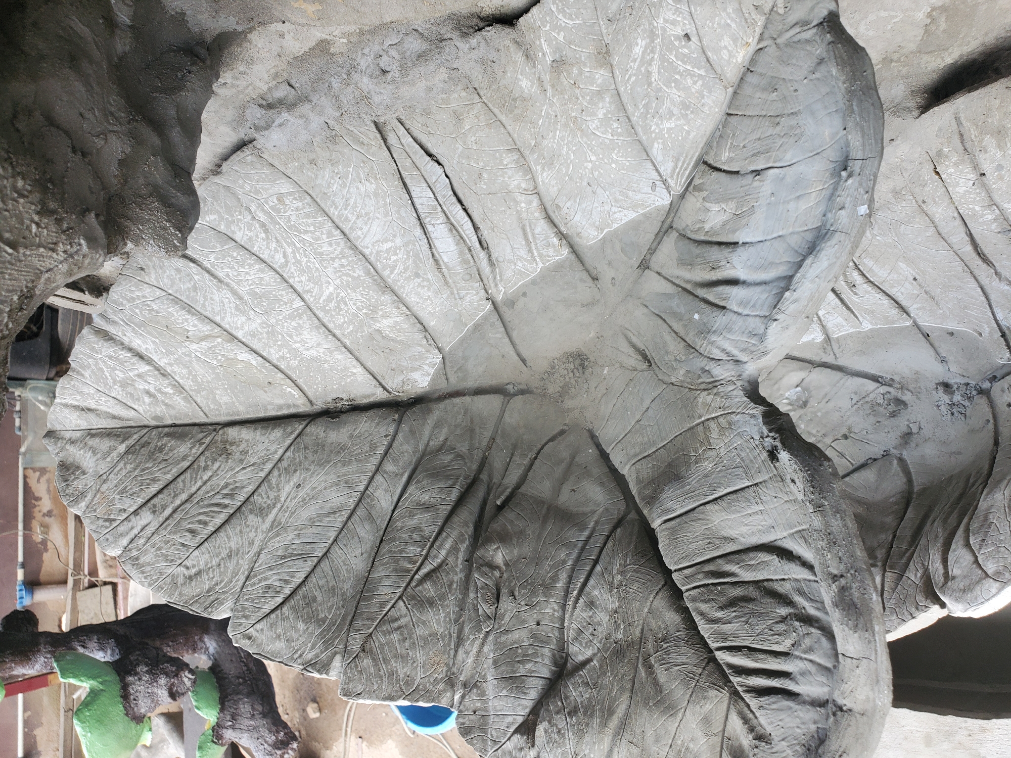 An overhead shot shows a large, gray, concrete sculpture of a leaf, resembling an elephant ear or taro leaf, with prominent veining. The leaf sculpture is wet in the center, suggesting it might be a water feature or has recently rained. To the left, a portion of another sculpture is visible, appearing to be a tree trunk with green leaf-like elements. The background suggests an outdoor workshop or construction area with sand, a blue bucket, pipes, and other construction materials. The overall scene is likely in Jos, Nigeria, given the context of the prompt. There are no people visible in the image.