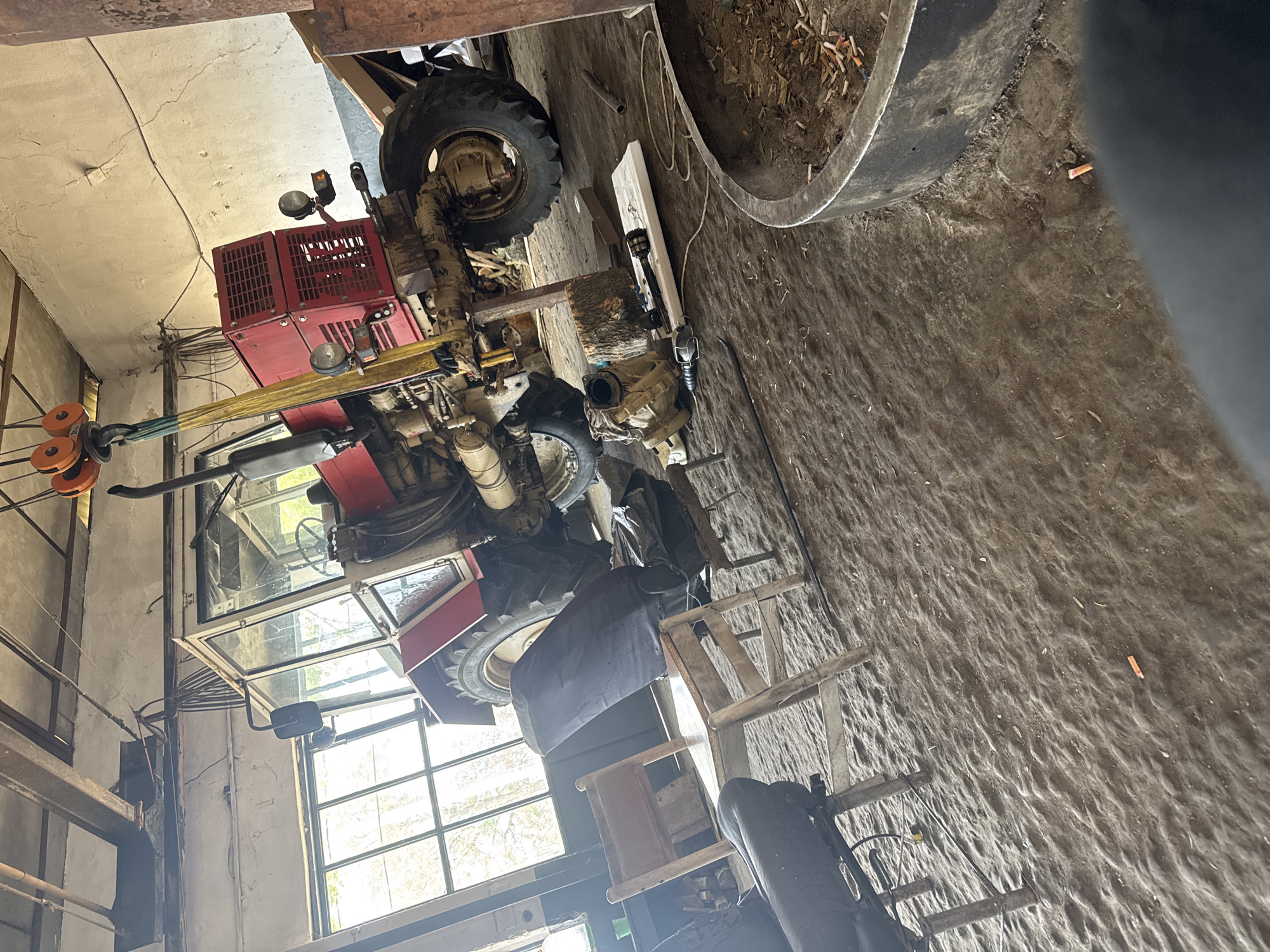 The image depicts an indoor scene within what appears to be a workshop, garage, or barn in Sarnevo, Bulgaria. The primary subject is a large, red tractor, suspended or propped up, suggesting it is undergoing maintenance or repair.
The tractor, partially disassembled, features a red chassis, a visible engine, large black tires, and a cab with glass windows. It is supported by yellow and green ratchet straps, with an orange pulley system visible on the left side of the frame, indicating a hoisting operation. The front of the tractor has multiple lights.
The floor is made of uneven dirt, scattered with various debris. To the right of the tractor, a large, dark metal ring or pipe, possibly a section of a culvert, is visible, filled with soil and numerous discarded cigarette butts. More cigarette butts are scattered on the dirt floor. Nearby, wooden planks, logs, and what appears to be a disassembled transmission or differential housing are placed on the ground. A small, rustic wooden stool or ladder is also present.
The walls are light-colored, possibly concrete or plaster, showing some cracking and exposed wiring. Overhead, wooden or metal beams are visible. Several large windows, some with gridded panes, allow natural daylight to illuminate the space, suggesting it is daytime. No people are visible in the scene, and there is no clearly legible text. The overall atmosphere is one of active work or storage in a rural or industrial setting.
