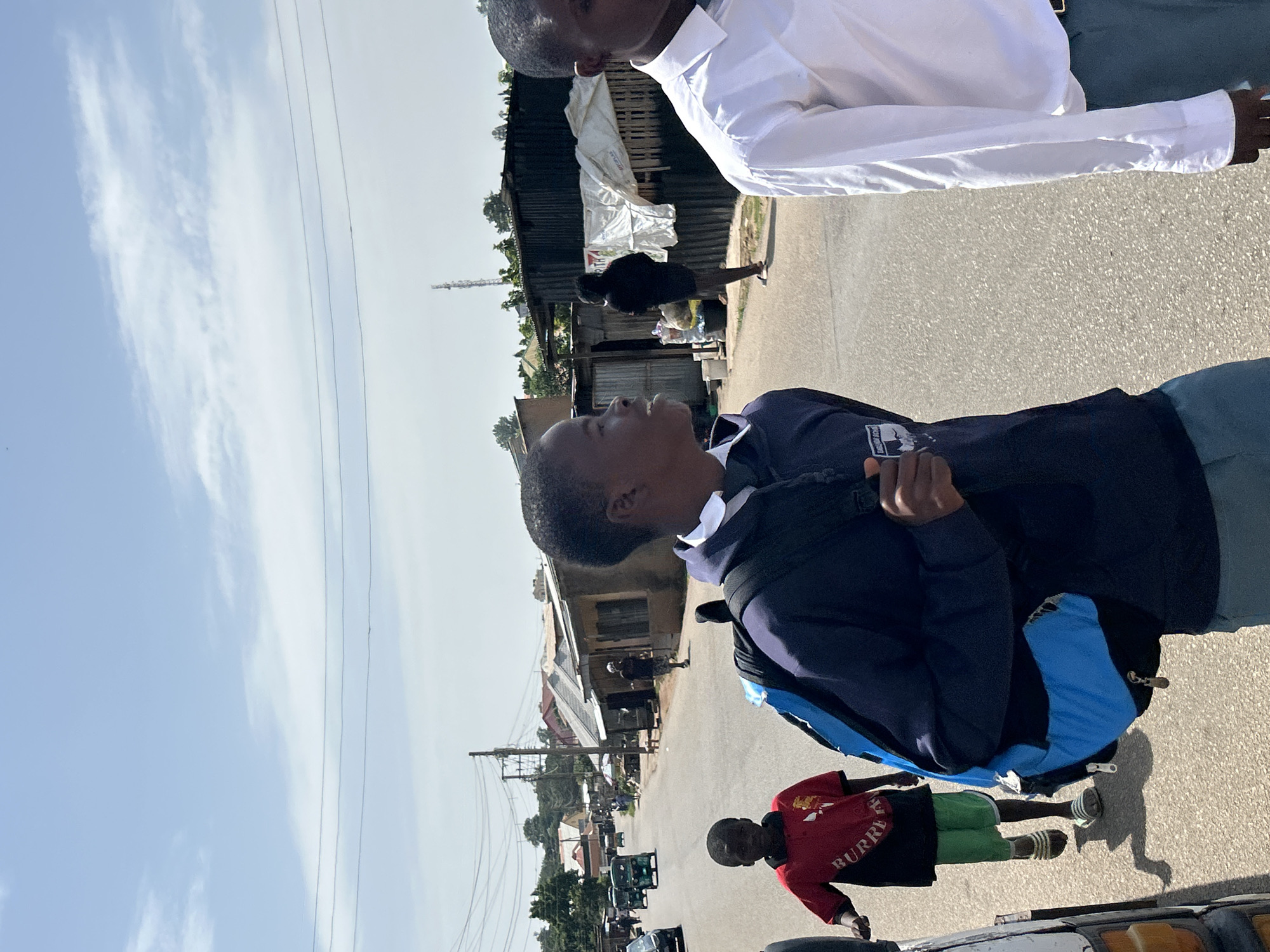 The image, taken in Jos, Nigeria, shows a street scene under a bright, partly cloudy sky. The sun appears to be high, suggesting it is midday. Several people are visible. In the foreground, a young person wearing a dark blue jacket over a collared shirt looks upwards and to the left, with a backpack over their shoulder. To their right, another young person in a red shirt with green shorts and striped sandals stands, facing towards the right. Behind them, a person in a white shirt is partially visible from the chest up, also looking away from the camera.
The background reveals a street lined with buildings, some appearing to be shops or informal market stalls with corrugated metal roofs and tarpaulin coverings. A telecommunications tower stands in the distance. Various vehicles, including what appear to be tricycles (popular in Nigeria as taxis) and cars, are present on the road, indicating active traffic. The overall atmosphere suggests a typical day in a bustling urban or semi-urban environment. The clothing of the individuals, particularly the school-like uniforms, hints at an educational setting or a group of students. The presence of utility poles and wires further establishes the urban context. The image is oriented vertically, with the top of the frame showing the sky and power lines.