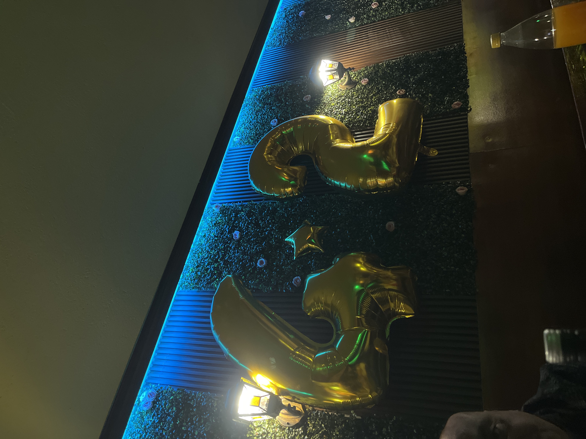 The image shows celebratory decorations in a dimly lit environment. Two large gold foil balloons, shaped like the numbers "2" and "5," are mounted on a textured wall adorned with artificial greenery and small rose accents. A small gold star-shaped balloon is positioned between the number balloons. A strip of blue LED lighting runs along the edge of the wall. Two small, modern, wall-mounted lights are positioned near the balloons, casting light. In the upper right corner, a partially visible plastic bottle containing orange liquid is present. A portion of a person's head is visible in the lower right corner. The location is Lozarevo, Bulgaria.