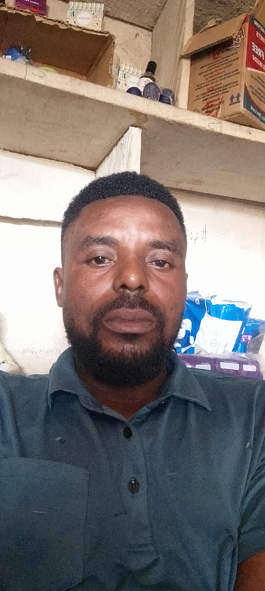 The image is a close-up, selfie-style portrait of a man with short, curly black hair and a beard, wearing a dark teal collared shirt. He is positioned in the foreground, with shelves filled with various items, including boxes and bottles, visible in the background. The lighting suggests an indoor setting. The man appears to be looking directly at the camera. The environment suggests a storage area or a shop. There are no visible indications of the city of Durumi, Nigeria, in the image itself.