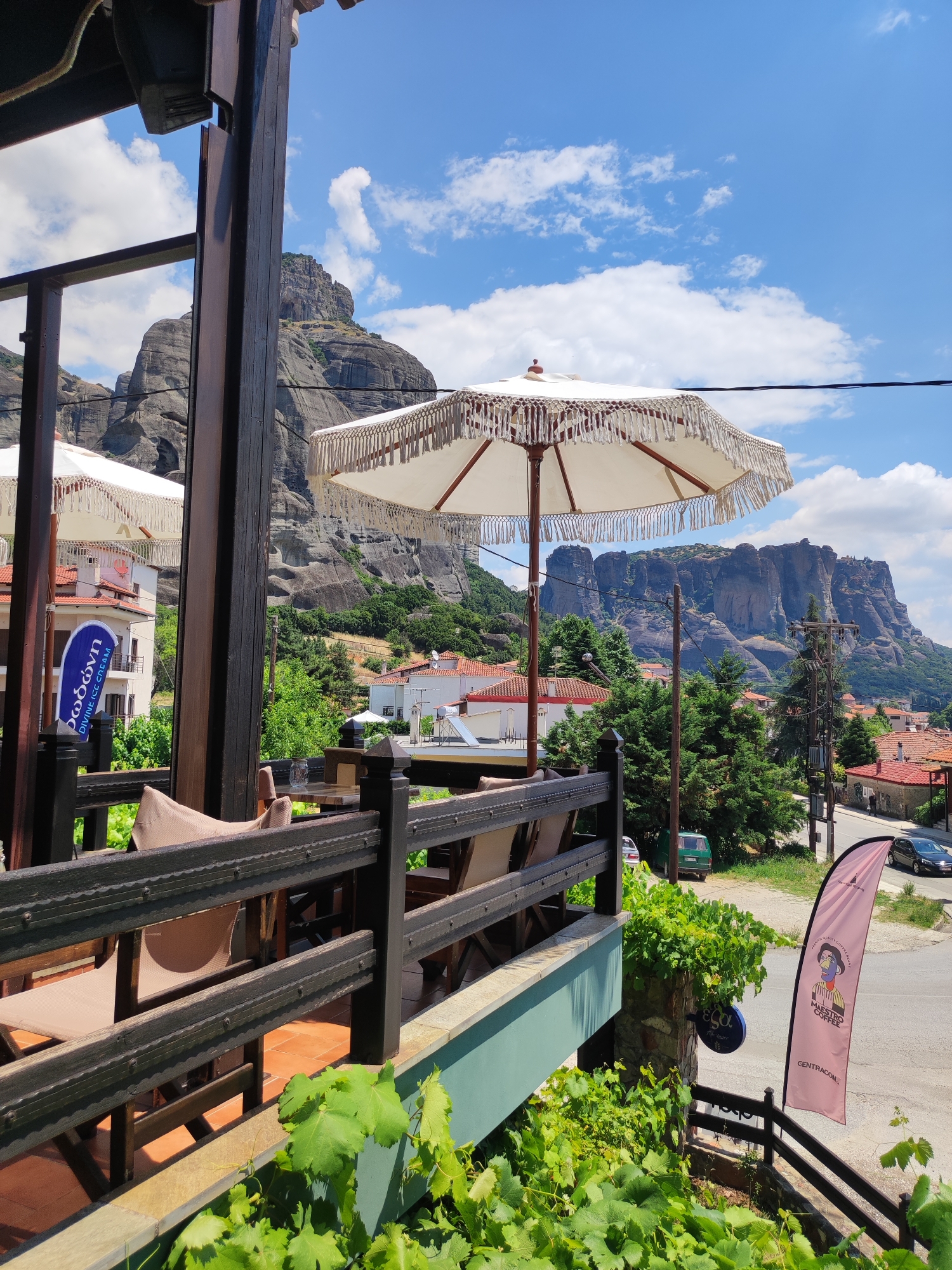 This is a vertical, eye-level shot taken on a sunny day in Kalabaka, Greece, offering a view from a restaurant or cafe's outdoor seating area towards a mountainous landscape and the town. In the foreground, a dark wooden railing encloses a patio with chairs and tables, some covered with tan fabric. Lush green vines grow in front of the railing. Two white, decorative umbrellas with fringed edges are prominent above the patio.
Beyond the patio, the town of Kalabaka is visible, with buildings featuring red-tiled roofs nestled among green trees. In the background, the impressive Meteora rock formations rise dramatically against a bright blue sky dotted with fluffy white clouds. A tall, dark wooden support pillar is on the left side of the frame. Various signs are visible, including a blue oval sign that reads "Δώσων DIVINE ICE CREAM" and a pink feather flag with a caricature and the text "MAESTRO COFFEE CENTRACOM". Cars are parked and driving on a road in the middle ground. The overall impression is one of a relaxed, scenic dining experience with a backdrop of natural and architectural beauty.