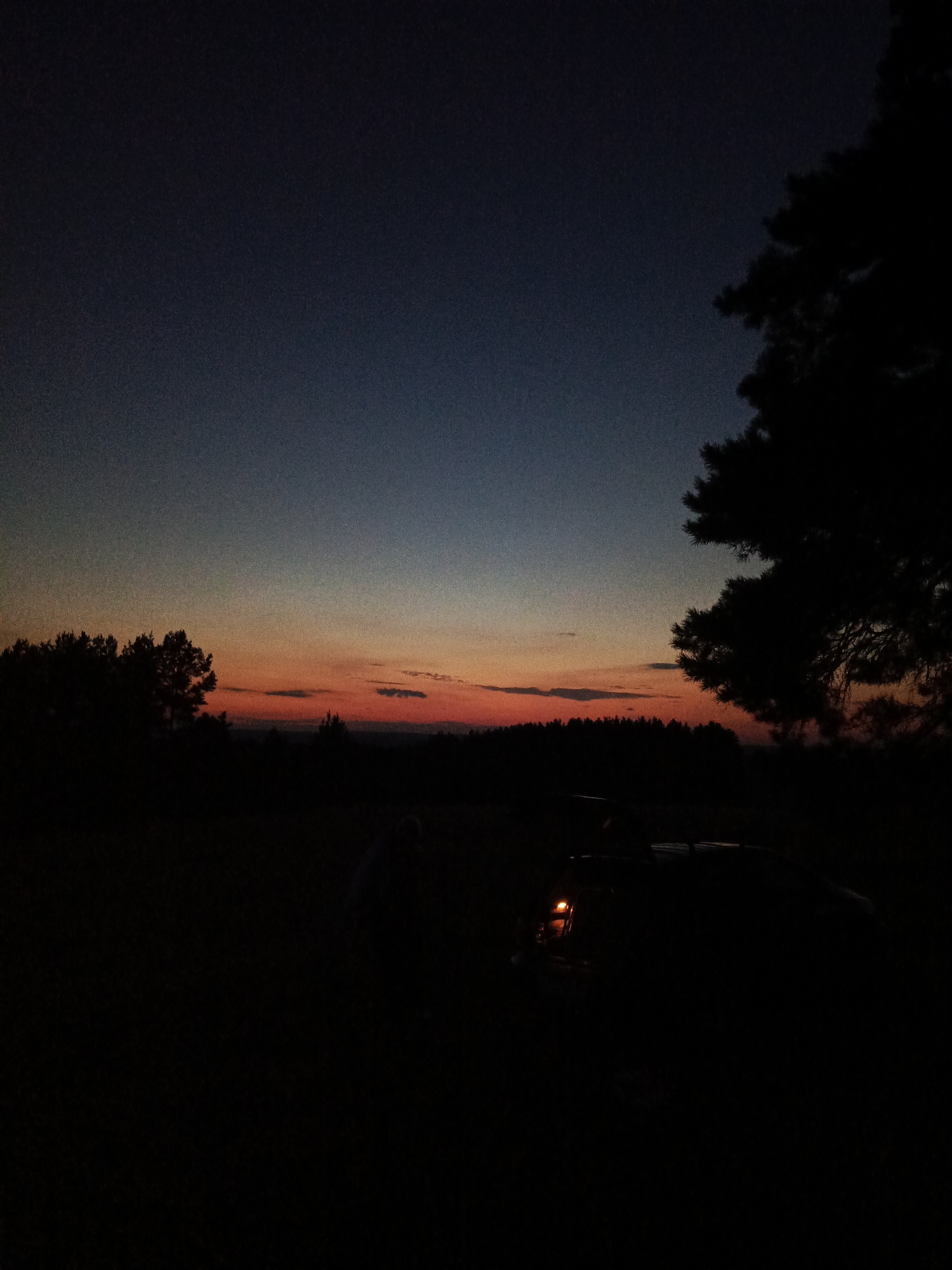 The image captures a sunset over a rural landscape, likely in or near the village of Chumna, Russia. The sky displays a gradient of colors, from a deep twilight blue at the top to shades of orange and red near the horizon, indicating the end of daylight. Silhouetted against the colorful sky are the dark outlines of trees on the left and a dense forest line in the background. In the foreground, a dark object, possibly a vehicle, is partially visible, with a single orange light illuminating the immediate surroundings. The overall scene is dimly lit, with a low level of detail in the foreground due to the fading light.
