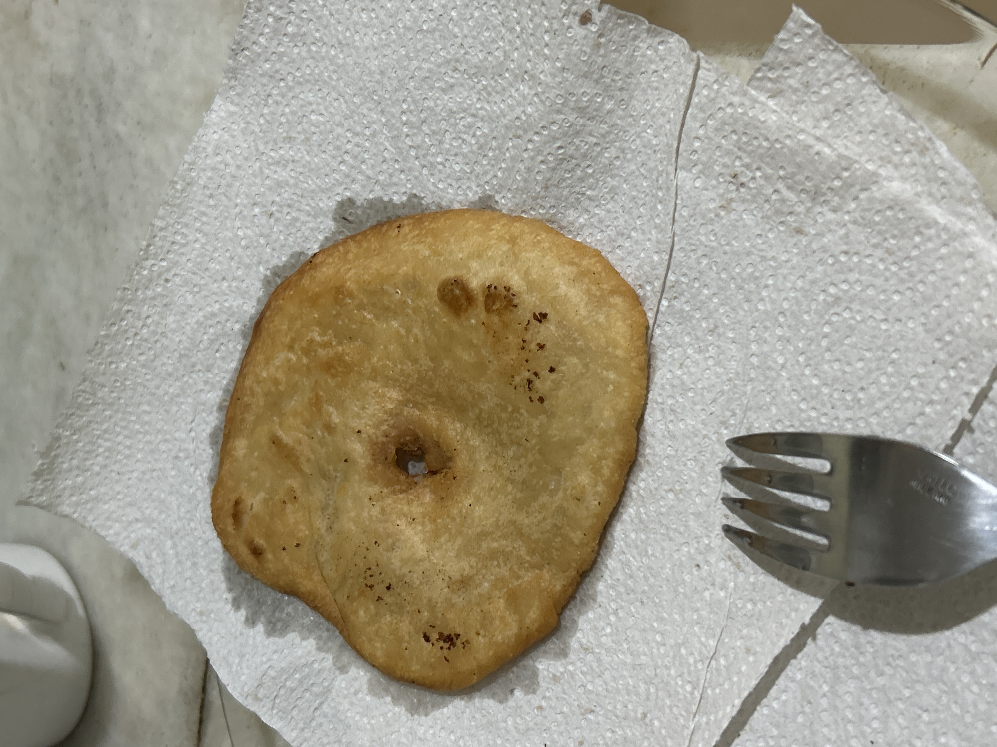 A fried pastry, possibly a sopaipilla or a similar fried dough, rests on a white paper napkin. The pastry is golden brown and has an irregular, somewhat circular shape with a hole in the center. There are small, darker brown spots on its surface, indicating where it may have fried more intensely. The napkin is textured and appears slightly greasy where it has absorbed oil from the pastry. To the lower left of the pastry, the tines of a silver fork are visible, casting a shadow on the napkin. In the upper left corner, out of focus, is the bottom part of a white ceramic mug or pitcher. The background is a neutral, possibly marble or tiled surface, suggesting an indoor setting. No specific location in Buenos Aires, Argentina, can be identified from the visual information provided.