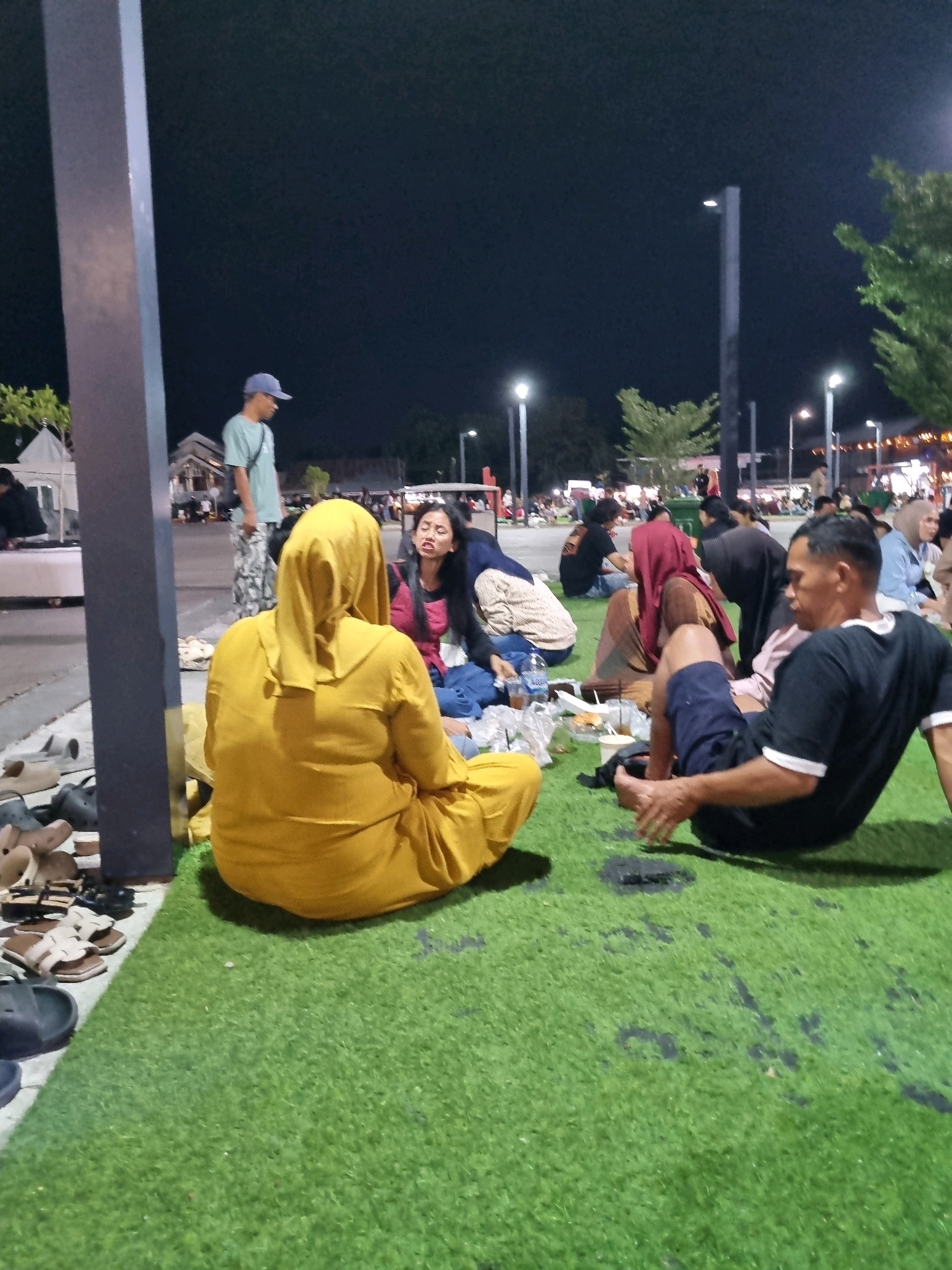 The image shows a nighttime scene at the Tanamodindi City Market in Palu, Indonesia. A group of people are gathered on artificial turf, with some seated and others talking. In the foreground, a person in a yellow hijab is seated facing away from the camera. To their right, a person in a black t-shirt and blue shorts is also seated. Behind them, other individuals are visible, including a woman in a pink top and a person wearing a red hijab. A man in a light blue t-shirt and a blue cap stands in the mid-ground. The market is illuminated by streetlights and lights from stalls, creating a lively atmosphere. Various footwear is scattered on the ground near a pillar in the left foreground.