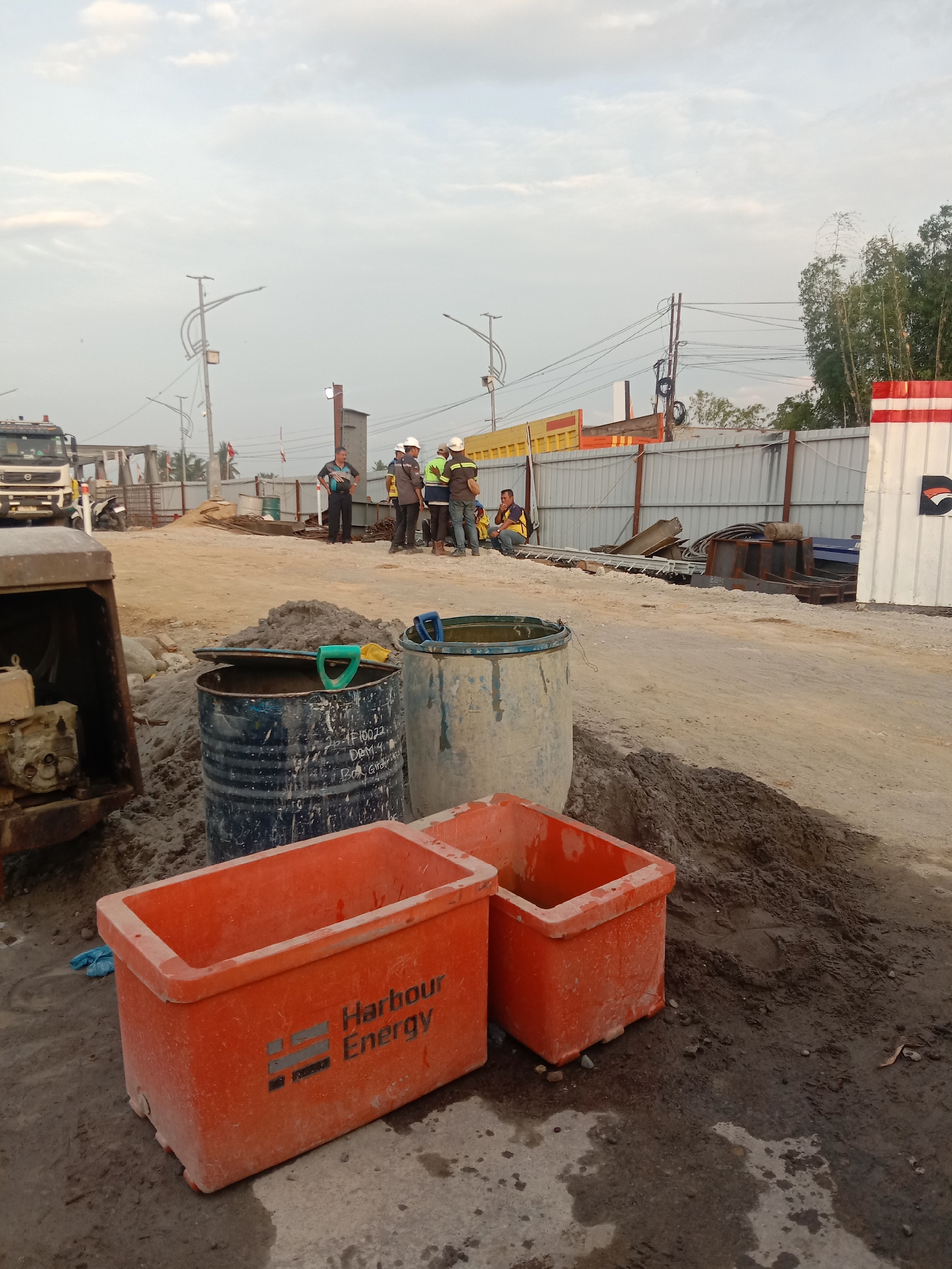 A group of four construction workers in hard hats and reflective vests stand and sit on a dirt path at a construction site in Lhoknga, Indonesia. In the foreground, two orange plastic bins with the "Harbour Energy" logo sit on the ground. Behind them, two large metal drums are visible, one blue and one light gray. A construction vehicle, possibly a truck, is parked on the left side of the image. The background shows a fence and various construction materials, with a cloudy sky above.