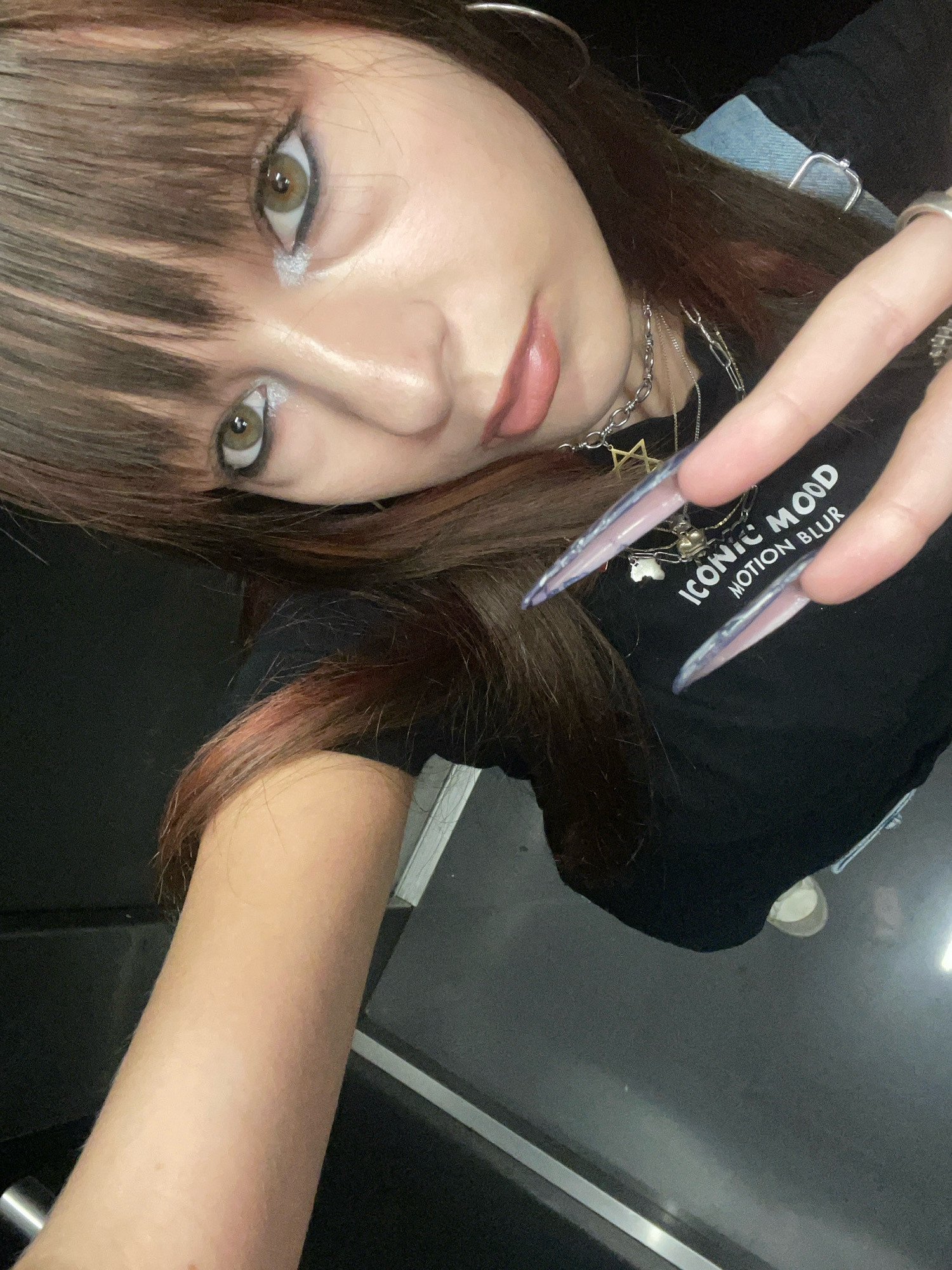 The image shows a close-up, tilted selfie of a young woman with long, dark brown hair and bangs. She is wearing a black t-shirt with white text that reads "ICONIC MOOD MOTION BLUR." She has dramatic eye makeup, including winged eyeliner and silver eyeshadow. Her lips are glossy and pink. She is wearing several silver necklaces, one with a star pendant and another with a small heart charm. Her fingernails are long, pointed, and appear to be artificial, with a gradient of purple and white. She is also wearing rings on her fingers. The background is dark and out of focus, suggesting an indoor setting. There is no discernible indication of location in the city of Raanana, Israel.