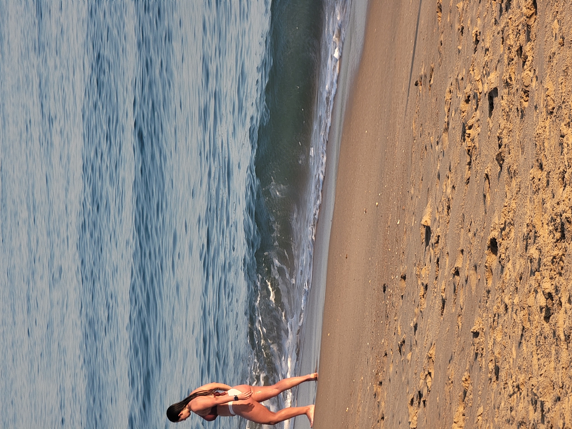 A woman wearing a brown bikini is walking along the shoreline of a beach in Quarteira, Portugal. She is facing left and her hair is dark and pulled back. The ocean water is a calm blue with small waves gently lapping the shore. The sand is light brown and has footprints and some small shells scattered across it. The scene is illuminated by natural sunlight, casting a warm glow on the sand.