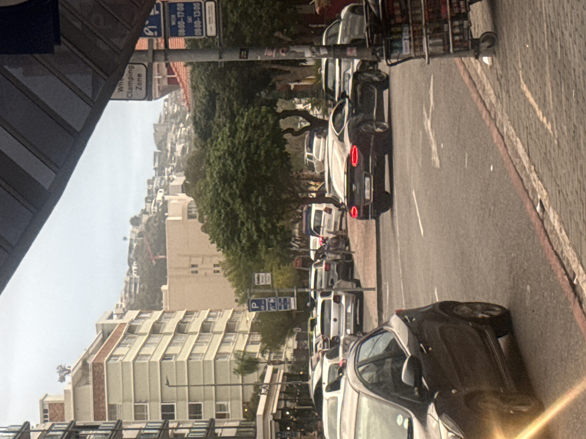 The image depicts a street scene in Cape Town, South Africa. A row of parked cars lines the right side of the road. A dark-colored car is parked closest to the viewer on the right side. On the left side of the street, a building with multiple floors is visible. Above the building, the landscape rises. There are street signs indicating "Wheel Clamping Zone" and parking regulations with times "08:00-17:00" on weekdays and "08:00-13:00" on Saturdays. The sky appears overcast.