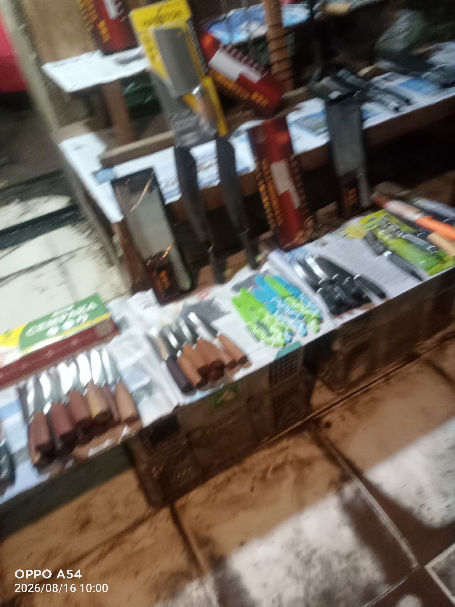The image depicts a market stall in Jambi, Indonesia, exhibiting a variety of knives and utensils. The stall is cluttered with items, including a green box with text in Indonesian, several knives with wooden handles arranged in rows, and packaged knives with colorful handles in blue, green, and black. In the background, there are shelves with more items, some of which appear to be tools or kitchenware. The overall impression is that of a bustling market environment, though the low resolution and blurriness of the image make it difficult to discern specific details about the people present or precise location within Jambi. The lighting suggests it might be indoors or an area with artificial light. The bottom left corner displays "OPPO A54 2026/08/16 10:00", indicating the device and date of capture.