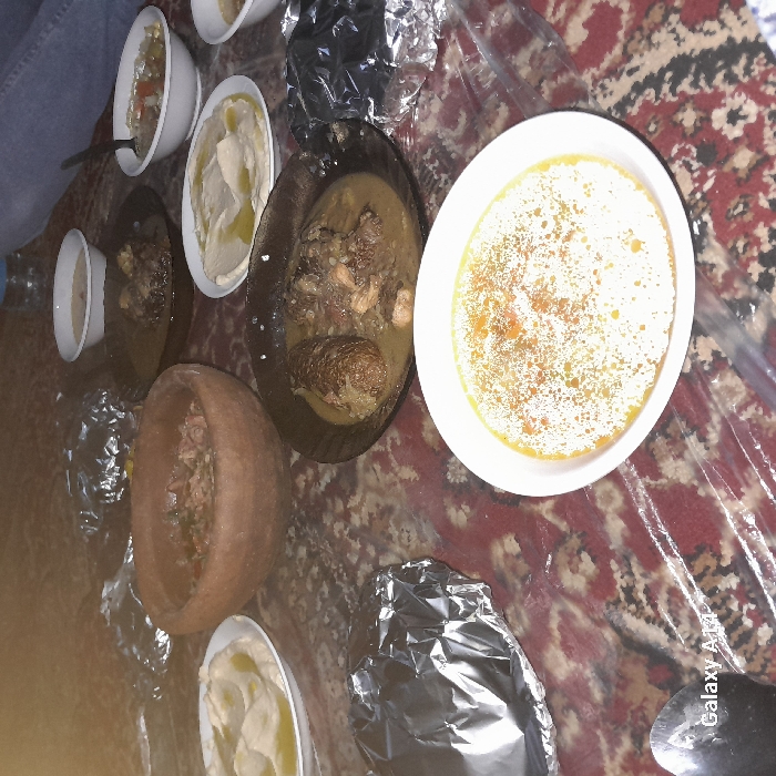 The image displays a spread of food laid out on a patterned rug, covered by a clear plastic sheet. The setting appears to be an indoor meal, likely eaten on the floor, a common practice in some cultures.
In the upper left corner, a person's leg clad in blue jeans is visible, suggesting someone is seated at the meal.
Several dishes are arranged across the rug:
* A large white oval bowl in the center-right holds a yellow or light orange soup or stew, possibly lentil soup, with visible reddish specks on its surface.
* Another white oval bowl, located towards the upper-middle left, contains a creamy, light-colored dip, most likely hummus, with a drizzle of olive oil on top.
* To the far upper-left, a smaller white bowl contains what appears to be a salad or a mixture of chopped vegetables. A spoon is visible within this bowl.
* Two dark, possibly black, shallow bowls are prominent in the middle. They both contain a rich, brownish-green meat stew with chunks of meat (possibly lamb or beef) and some larger, darker pieces that might be stuffed vegetables (like zucchini or eggplant) or substantial meat portions.
* A rustic, reddish-brown, possibly ceramic or clay, oval dish is situated in the lower-middle left. This dish also contains a stew or casserole, with visible ingredients and liquid.
* Several items are wrapped in aluminum foil, scattered around the food. A large, prominent foil-wrapped item is in the lower right corner, suggesting food that is either being kept warm or was transported in that wrapping.
* A small white bowl, possibly containing another dip or side, is partially visible near the blue jeans on the far left.
A black plastic bag is visible near the top center, next to some foil. The rug beneath the plastic sheet features a traditional pattern with shades of red, beige, and darker accents.
The overall scene depicts a communal meal, likely a family gathering or a shared dinner, with a variety of dishes suggesting Middle Eastern or Mediterranean cuisine.
In the bottom right corner of the image, the text "Galaxy A12" is faintly visible, indicating the device used to capture the photograph.