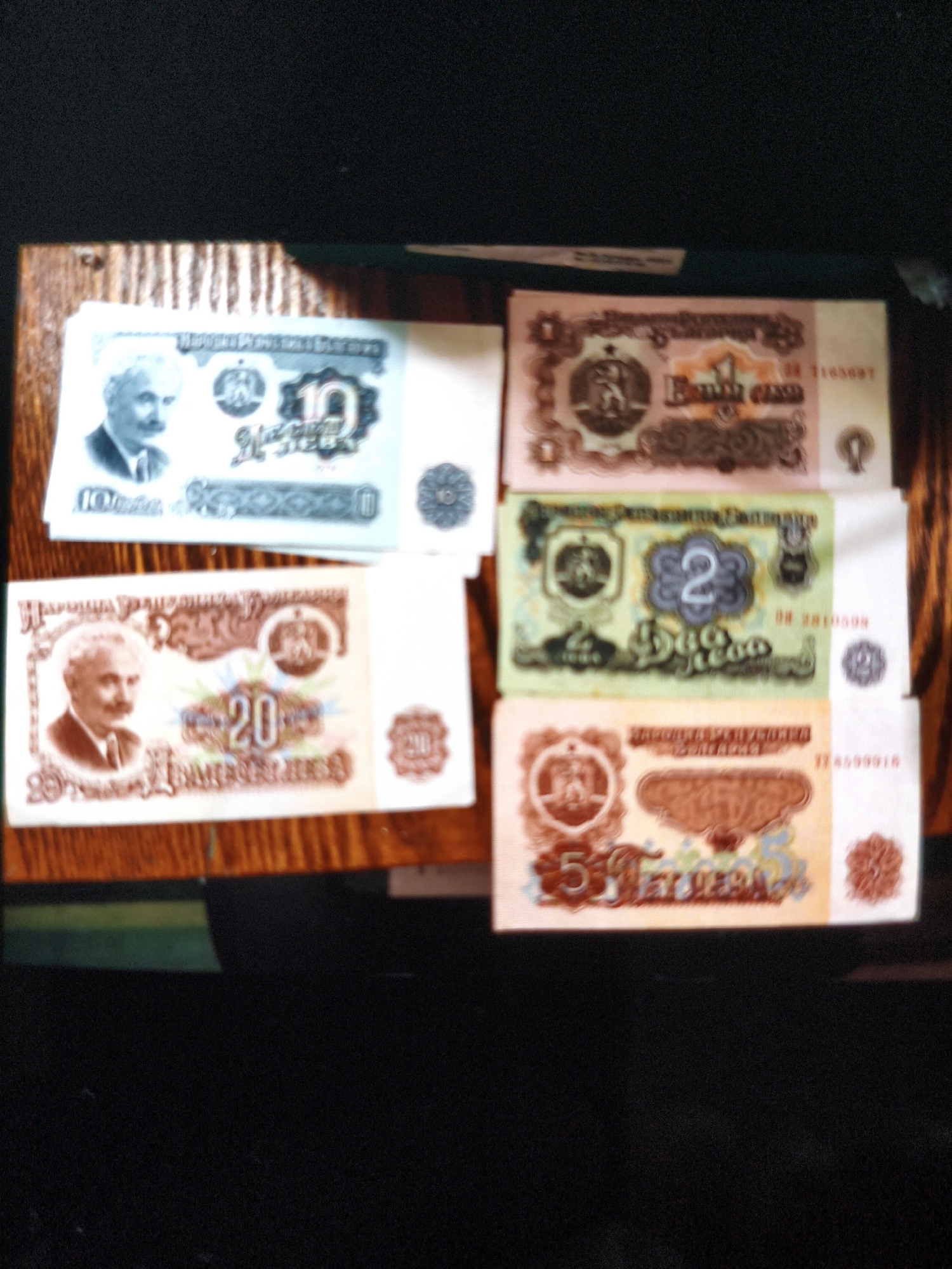 The image shows a close-up view of five Albanian banknotes of different denominations lying on a wooden surface. Clockwise from the top left, there is a 10 Lekë note with the portrait of Naim Frashëri, followed by a 1 Lekë note, a 2 Lekë note, a 5 Lekë note, and a 20 Lekë note with the portrait of Naim Frashëri. The notes are of various colors and designs. The wooden surface appears to be a table. The location in the city is unknown, unknown.