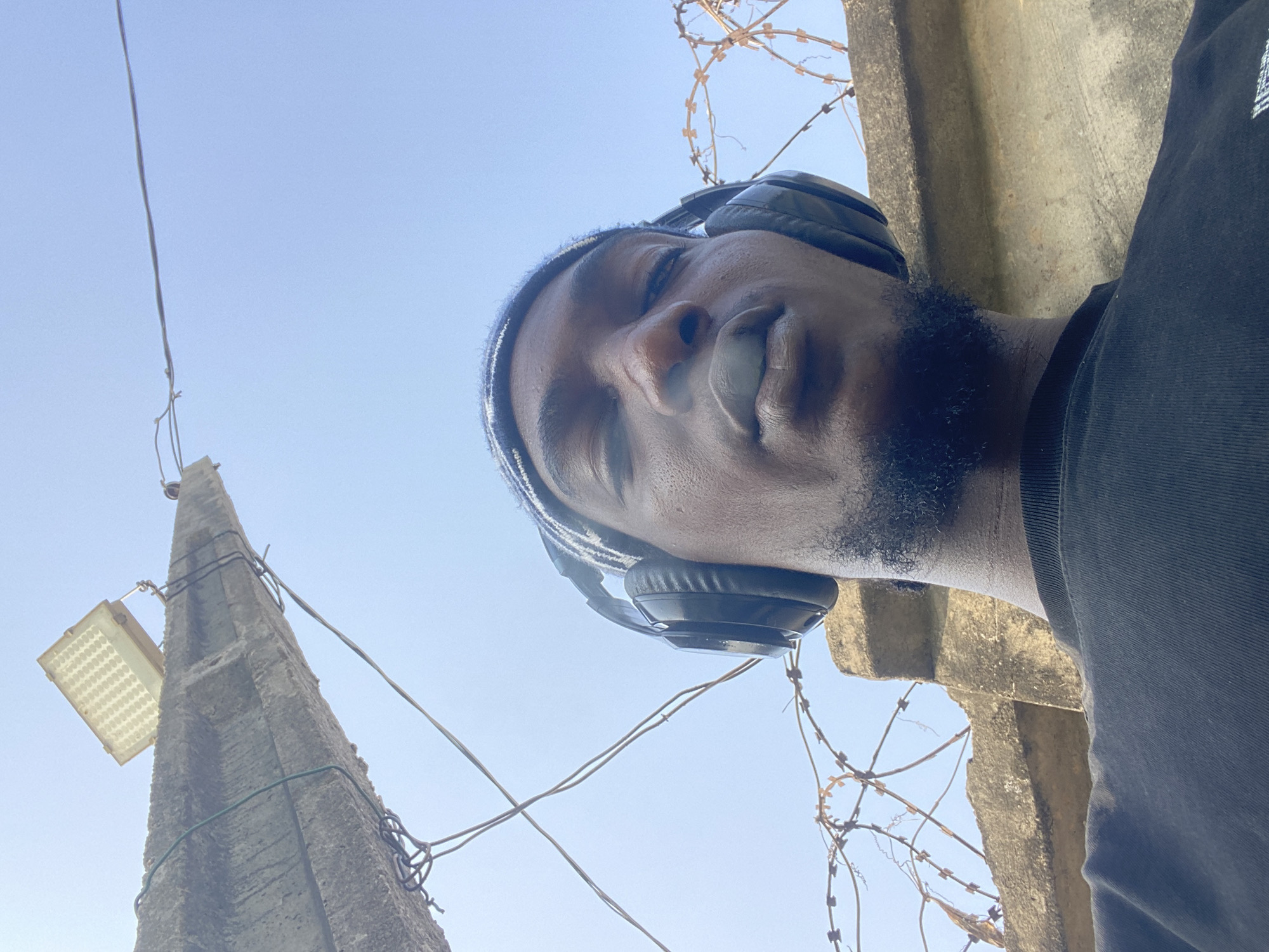 A low-angle shot shows a Black male wearing black headphones and a dark t-shirt. He has a short beard and appears to be chewing gum. Behind him, against a light blue sky, is a concrete structure, possibly a pole, with wires attached and a light fixture. Barbed wire is visible near the pole. The location is Lagos, Nigeria.