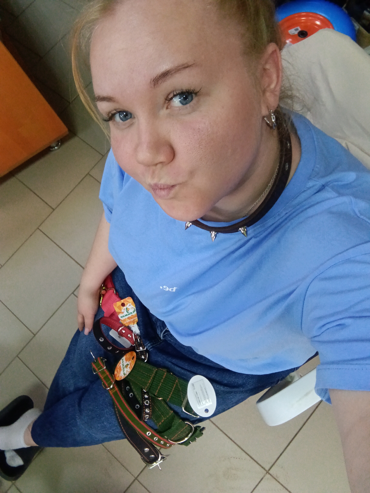 A woman is posing for a selfie indoors. She has light skin, blonde hair pulled back, blue eyes, and is wearing a light blue t-shirt, blue jeans, and a spiked collar. A variety of dog collars and accessories are draped over her lap. She has a small silver earring in her left ear and is pursing her lips for the photo. She is wearing black shoes with white socks. The background shows a tiled floor and a glimpse of furniture. The location is Фоки, Russia.