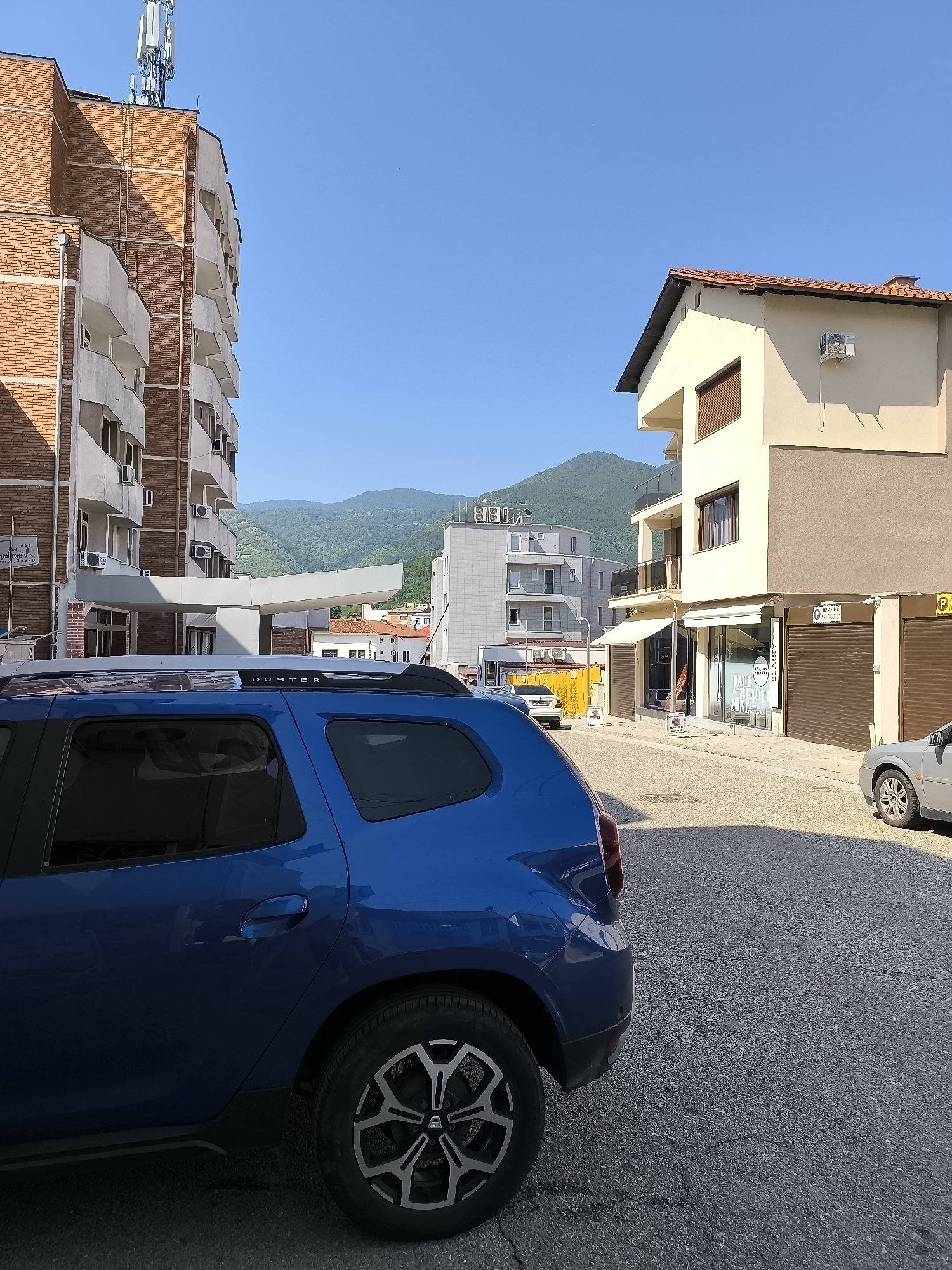 This is a street-level, eye-level, full-frame, vertical, exterior, daytime, medium-shot photograph taken in Gotse Delchev, Bulgaria. The scene features a blue SUV parked on the right side of the frame, partially obscuring a light-colored building with a red-tiled roof and a beige facade on the right. In the background, a multi-story brick building with white balconies is visible on the left, topped with telecommunications equipment. Beyond these structures, a range of green, tree-covered hills or mountains forms the backdrop under a clear blue sky. The street appears to be paved with asphalt, and several other vehicles, including a silver sedan, are parked or driving in the scene. The overall impression is of a sunny day in a European town, possibly during summer.