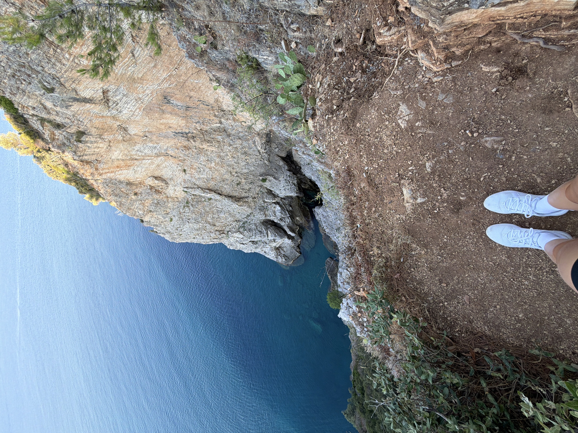 An aerial view shows the turquoise waters of the Adriatic Sea next to a steep, rocky cliff face covered in sparse vegetation. The cliff is composed of layered rock formations in shades of beige and tan, with some reddish-brown streaks. The water appears deep and clear, with darker patches indicating greater depth. On the edge of the cliff, a person's legs are visible, wearing white sneakers and dark shorts. The ground where the person stands is covered in dirt, small stones, and dry grass. A prickly pear cactus is growing on the cliff face. The scene suggests a natural, rugged landscape, likely in a coastal area.