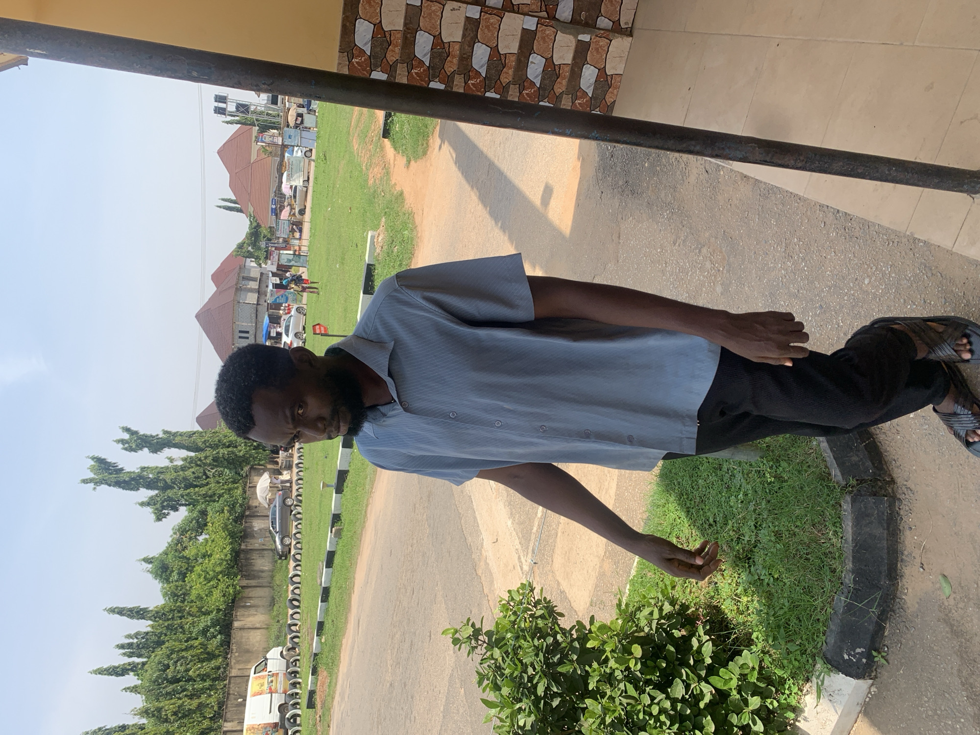 A dark-skinned man with a beard and short black hair, wearing a light blue short-sleeved collared shirt and black trousers, stands on a paved walkway next to a grassy area with a curb. He is facing slightly away from the camera, looking to his left. His left arm is bent at the elbow and his hand is near his chest, while his right arm is extended downward. In the background, across a road and grassy field, there are buildings with red tile roofs, trees, and several cars and a white van. The setting appears to be an outdoor environment with a clear sky.