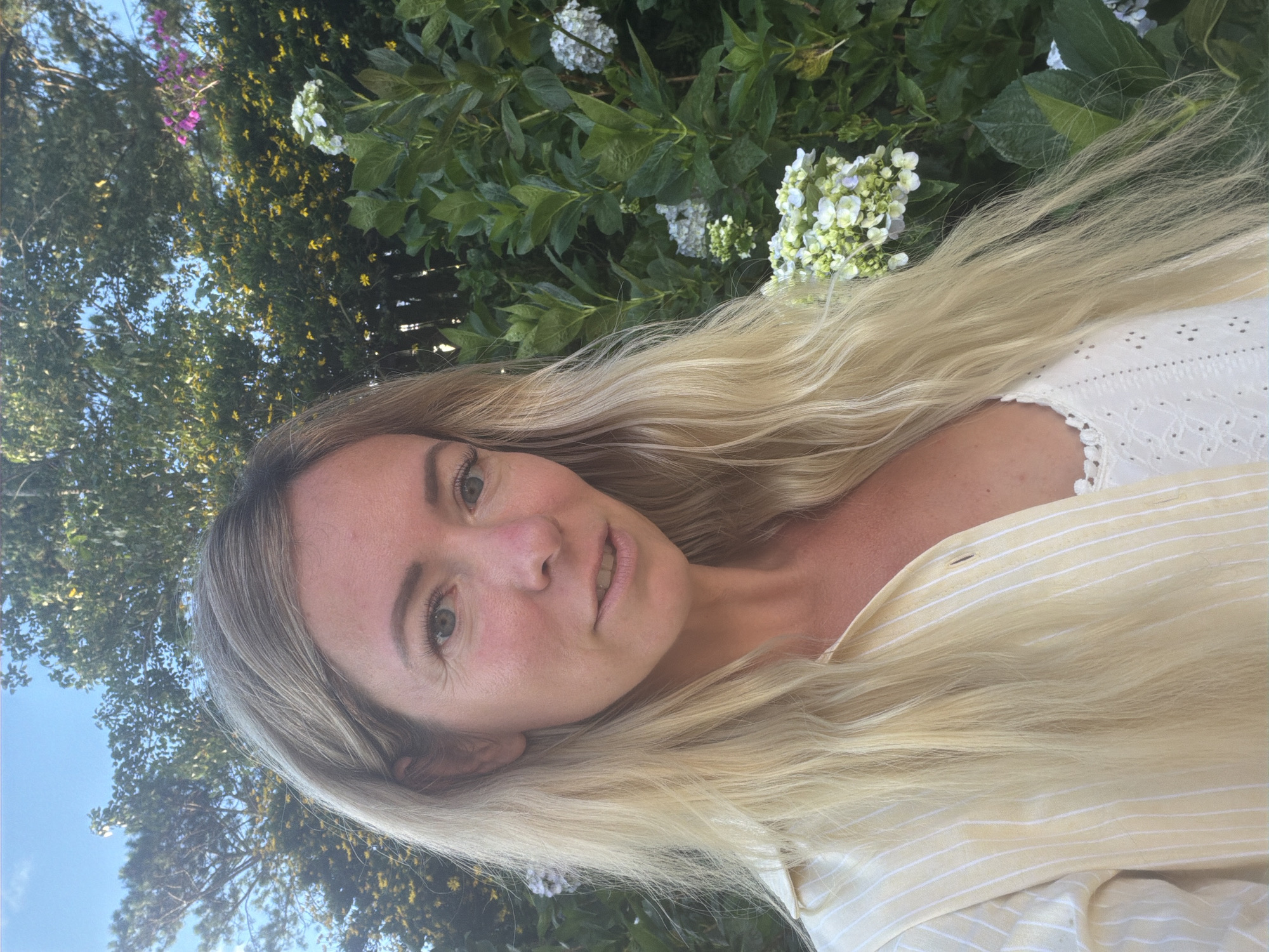 In the city of Xuan Thanh, Vietnam, a fair-skinned woman with long blonde hair and light green eyes is photographed from the chest up. She is wearing a pale yellow striped button-down shirt over a white lace camisole. The background is filled with lush green foliage and flowering plants, including white hydrangeas and yellow flowers, under a clear blue sky. The woman is looking towards the camera with a neutral expression. The lighting suggests it is daytime, possibly late afternoon due to the warm tones.
