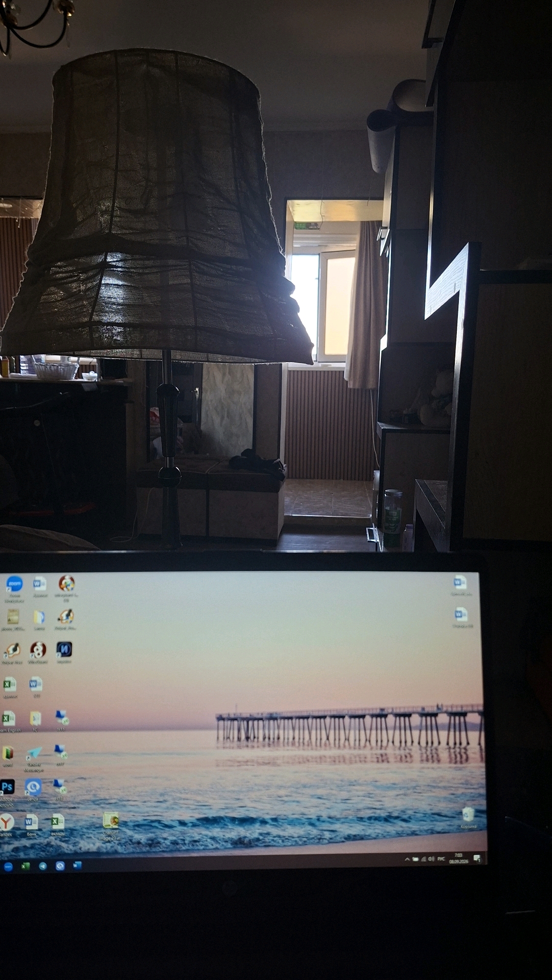 The image shows a close-up view of a laptop screen displaying a desktop wallpaper of a pier over a body of water. In the background, partially out of focus, is an interior scene featuring a large lampshade hanging from the ceiling. The room appears dimly lit, with a window visible in the distance that lets in natural light. The overall impression is that of a person using a laptop in a domestic or office setting. There is no indication of the city of Navoiy City, Uzbekistan, or any specific location.