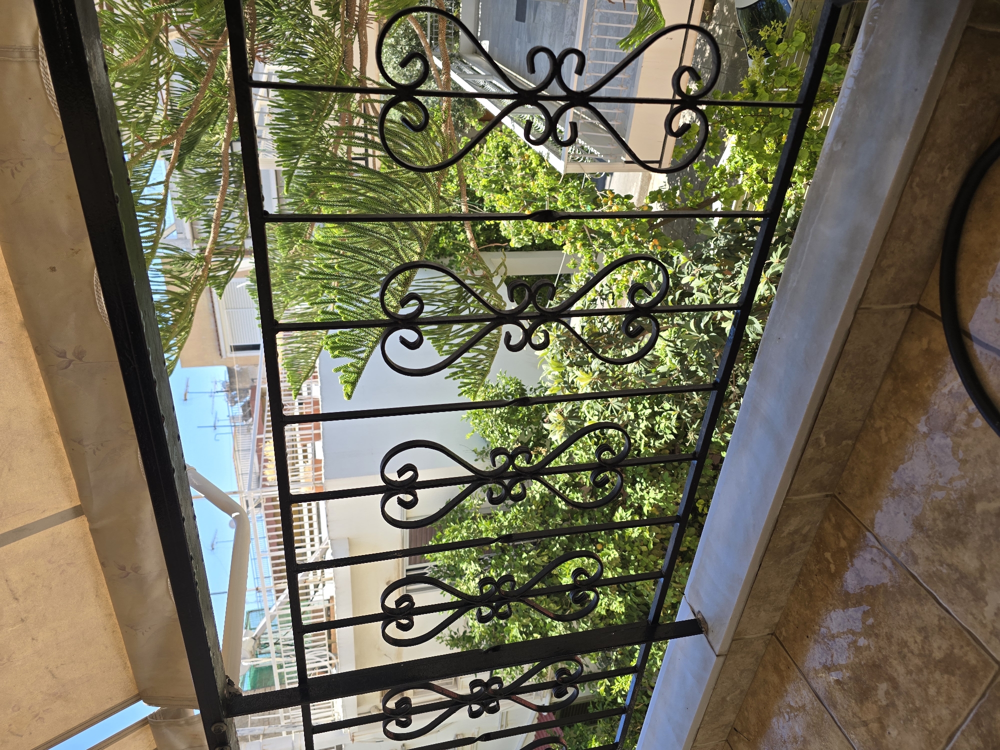 The image shows a view from a balcony in Ilion, Greece. The balcony has a light-colored tiled floor and a low, light-colored tiled wall. A black metal railing with ornate heart-shaped designs runs along the front of the balcony, partially obscuring the view of the surroundings. Beyond the railing, there is lush green vegetation, including a large conifer tree with needle-like leaves. In the background, several buildings are visible, some with white walls and balconies. The sky is blue, suggesting it is daytime. The scene appears to be residential.