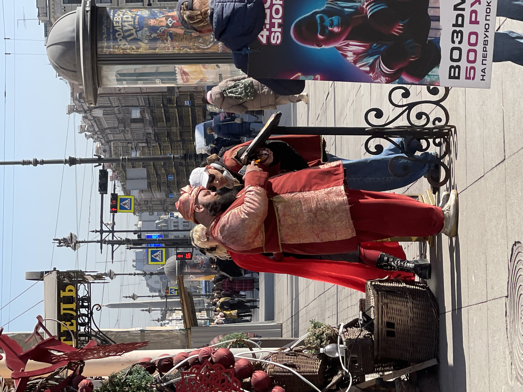 The image captures a scene in Saint Petersburg, Russia, where a person dressed in a theatrical costume, possibly for performance or entertainment purposes, interacts with another individual. The person in costume, adorned in a red and gold outfit with a matching turban, is holding a prop sword. They are standing on a sidewalk next to a decorative wrought iron fence. Behind them, other people can be seen walking along the busy street. There is a sign advertising "ЦРК С 37 ЯНВАРЯ," suggesting a circus or theatrical performance. A sign in the background reads "ВОЗЬМ 500 PY" which may indicate a promotional offer or price. To the left, there are red decorative elements and what appears to be a wicker basket on a small display. The urban backdrop includes buildings, traffic lights, and street signs, all under a clear sky.