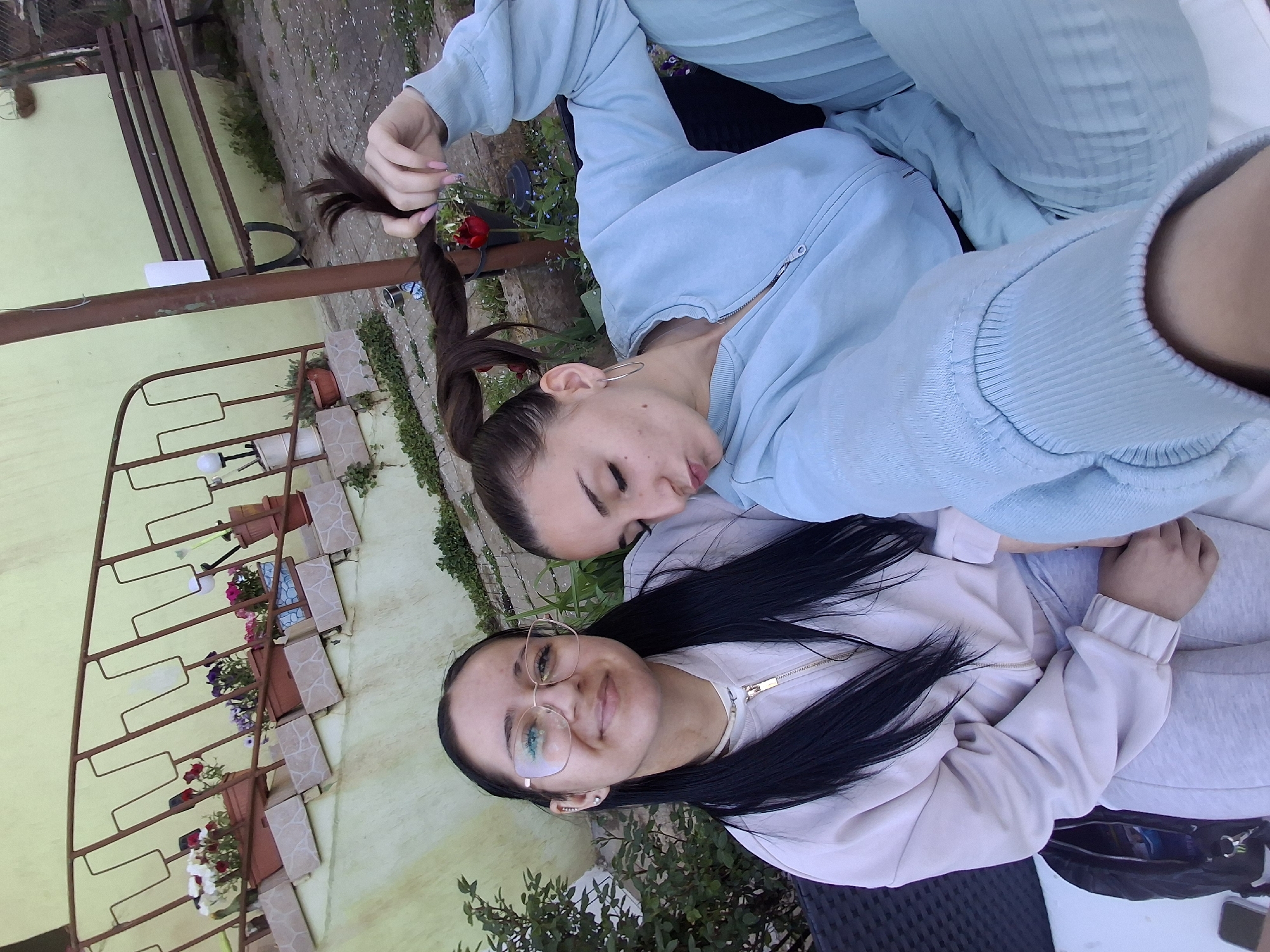 The image depicts two young women outdoors. The woman on the bottom has long, black hair and is wearing glasses and a light pink jacket. She is smiling. The other woman, who is on top of her, has brown hair pulled into a ponytail with a strand held in her hand and is wearing a light blue zip-up sweatshirt. They are in a garden setting, as evidenced by potted plants, greenery, and a metal trellis in the background. It also looks like they are located near a bench. It is unclear if the image was taken in Buhovo, Bulgaria.