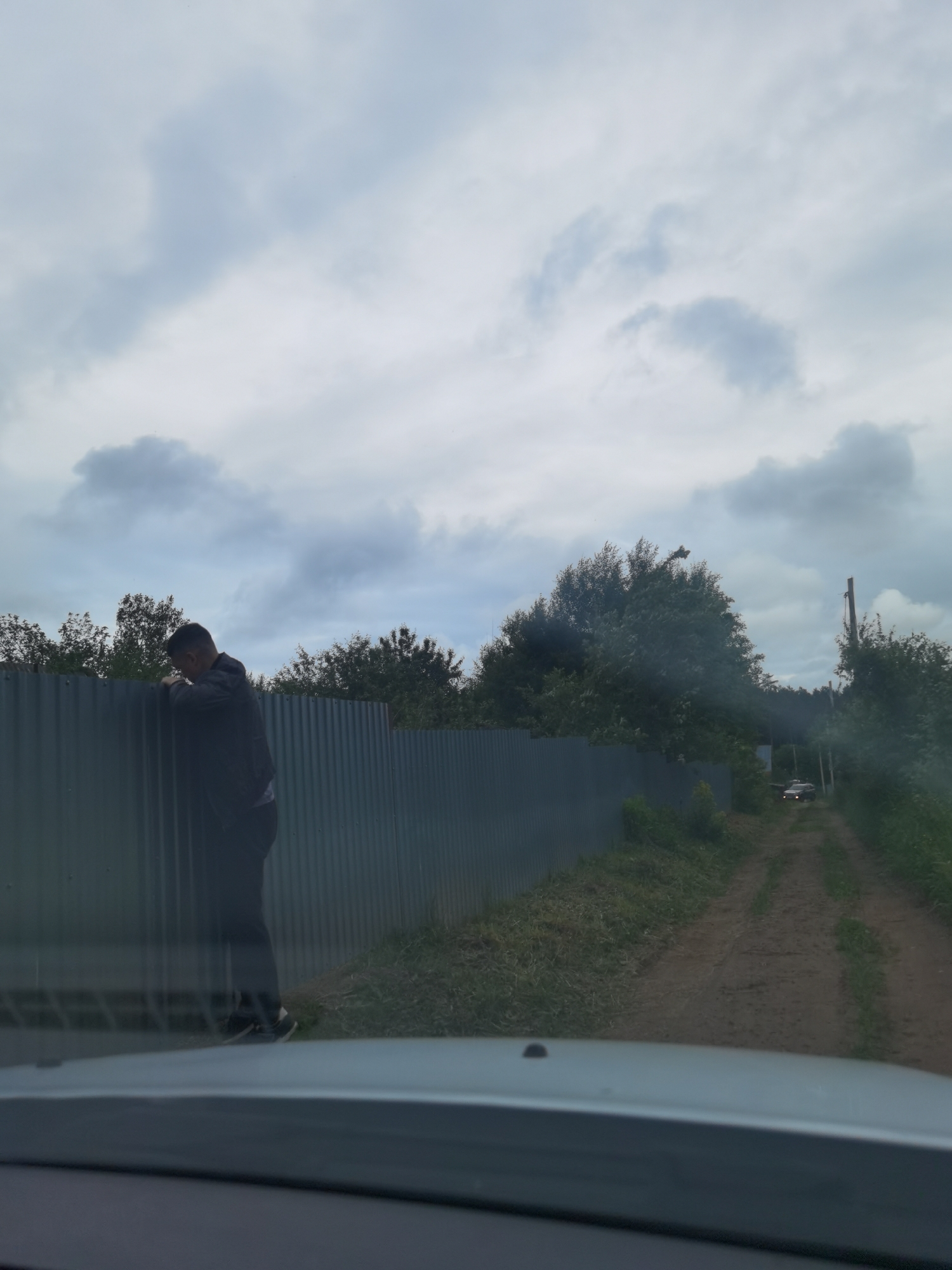 This image, captured in Прикамский, Russia, depicts a rural or semi-rural scene viewed from inside a vehicle, with the dashboard and windshield frame visible in the foreground. A cloudy, overcast sky occupies the upper two-thirds of the frame. In the mid-ground, a tall, dark grey, corrugated metal fence stretches horizontally. A male individual, dressed in dark clothing and sneakers, stands on the left side, leaning against the fence with his arms resting on its top edge and his head bowed. To the right of the fence, a dirt road with visible tire tracks extends into the distance, flanked by dense green trees and vegetation. A dark-colored car is faintly visible on the distant dirt road, and a wooden utility pole is present on the right side, partially obscured by trees.