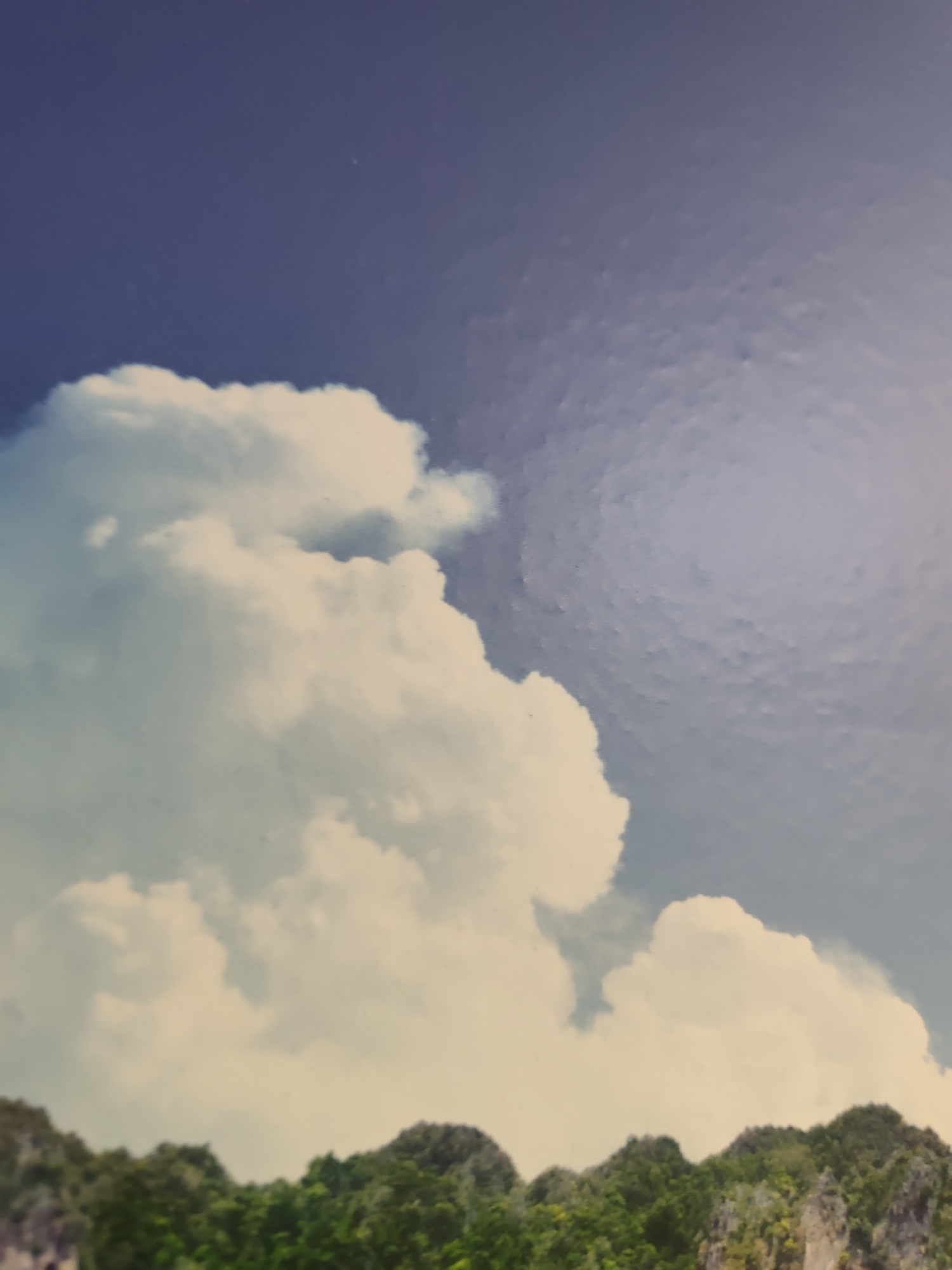 This image depicts a natural landscape scene located in the city of Pazardzhik, Bulgaria. The upper two-thirds of the frame are dominated by a sky displaying a transition from a deep blue on the far left to a lighter, somewhat mottled blue on the right. Large, voluminous white cumulus clouds are prominently featured on the left and central portions of the sky, with smaller, more diffuse clouds visible towards the right. The bottom third of the image shows a line of densely forested mountains or hills, characterized by lush green foliage covering irregular, rocky outcrops. The terrain appears rugged with visible rock formations, particularly on the right side. The scene is illuminated by natural daylight, and no people, animals, or man-made objects are visible within the frame.