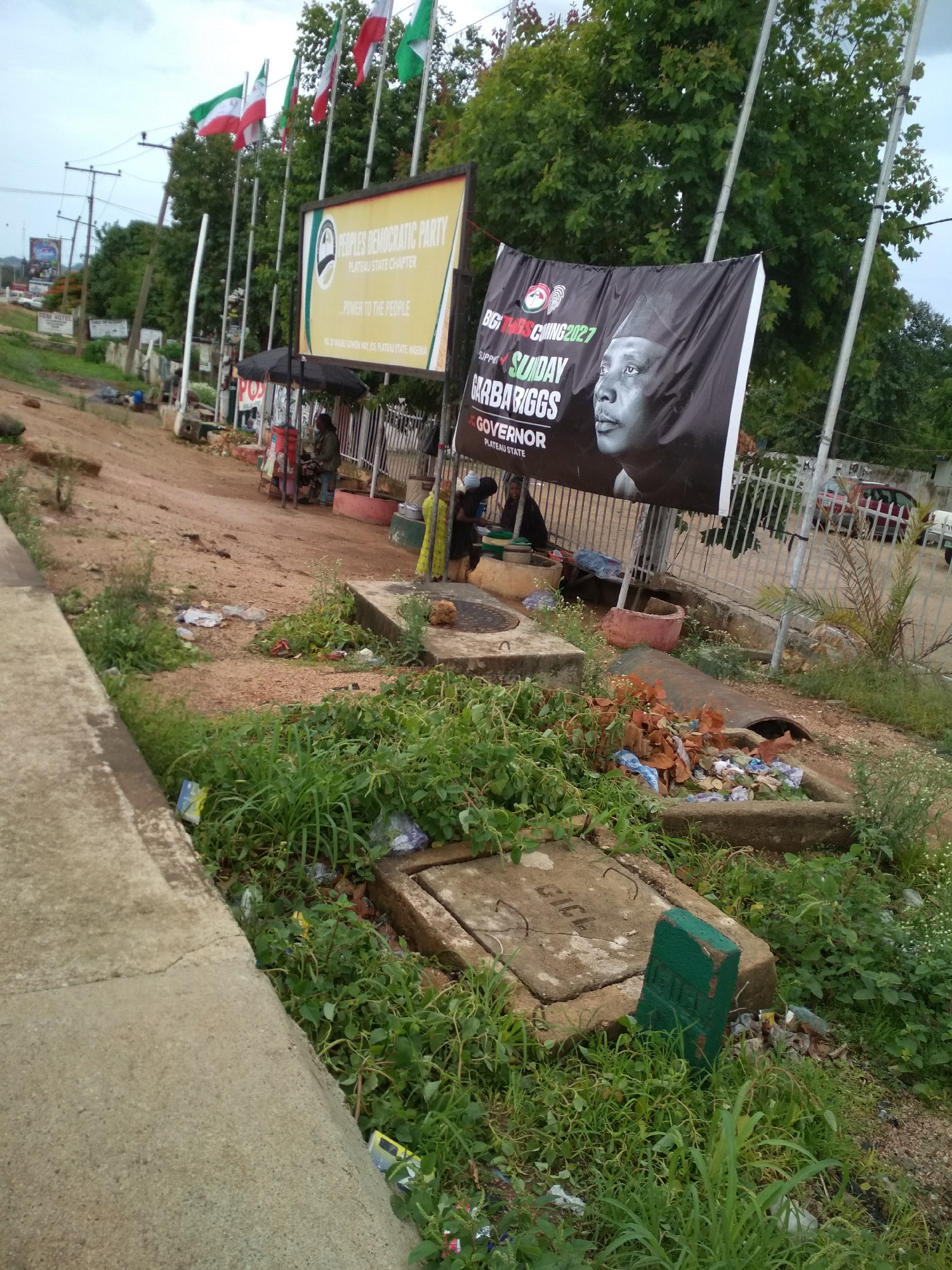 This media file presents an outdoor street scene in Jos, Plateau State, Nigeria, under an overcast sky. The foreground shows a concrete curb alongside an unkempt dirt strip densely covered with green grass and weeds, where numerous pieces of litter, including plastic bags and other debris, are scattered. Several concrete drainage covers or manholes are visible within this strip, one of which has "GIC" inscribed on it, and another is marked by a green painted block.
A reddish-brown dirt path extends into the mid-ground, leading past small, makeshift market stalls with umbrellas where a few individuals are present, possibly engaged in selling or daily activities.
In the background, two prominent political banners are displayed. One is a yellow billboard for the "PEOPLES DEMOCRATIC PARTY PLATEAU STATE CHAPTER," proclaiming "POWER TO THE PEOPLE" and specifying "JOS, PLATEAU STATE, NIGERIA." Adjacent to it is a large black banner promoting "SUNDAY GARBARIGGS GOVERNOR PLATEAU STATE" with "BIG THINGS COMING 2027," featuring a portrait of the candidate.
Behind the banners, multiple flagpoles fly flags, including what appear to be Nigerian national flags (green, white, green) and possibly party flags (red, white, green). A dense canopy of green trees forms a natural backdrop. Utility poles and lines are visible on the left, and in the distance, a sign for "FEM HOTEL" can be discerned. A metal fence separates the immediate area from a paved road where a red car and other vehicles are visible. The scene depicts a bustling roadside environment with ongoing political campaigning, daily commerce, and noticeable environmental neglect due to litter.