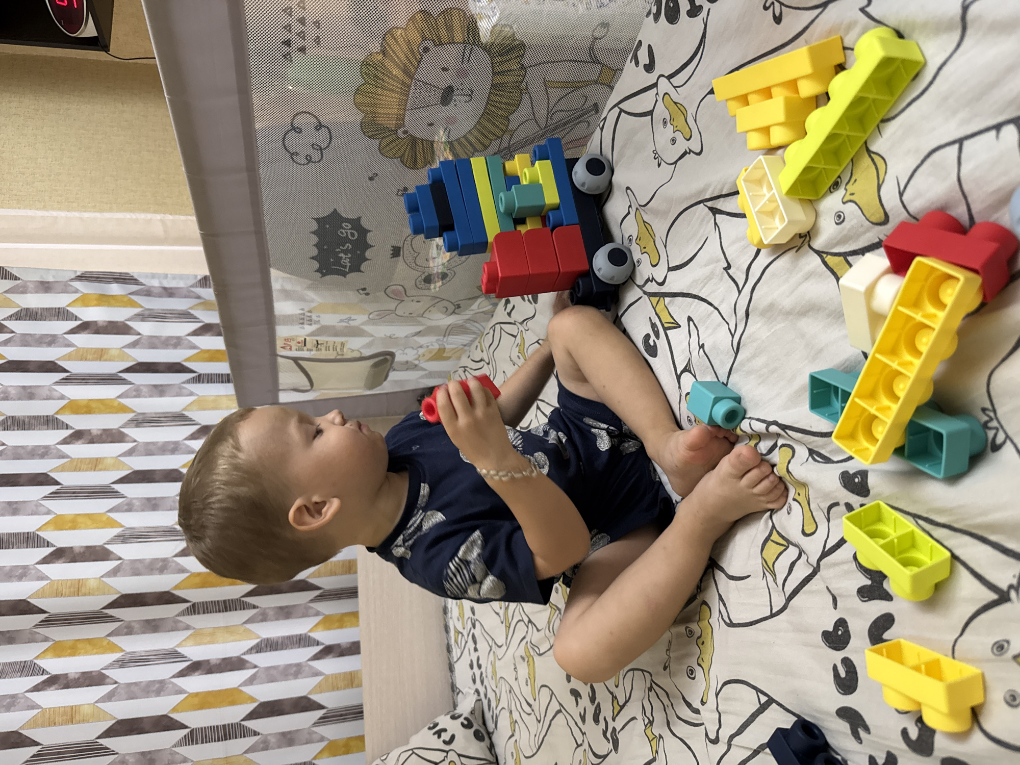 A young boy sits on a bed, playing with colorful building blocks. The boy is wearing a dark blue t-shirt with a pattern and dark blue shorts. He is holding a red block in his right hand. In front of him, a toy train made of blocks is assembled. The bed is covered with a white sheet featuring a pattern of ducks. In the background, a crib with a net is visible, decorated with cartoon animals and the text "Let's go". A digital clock with red digits is mounted on the wall above the crib. The room appears to be a child's bedroom.