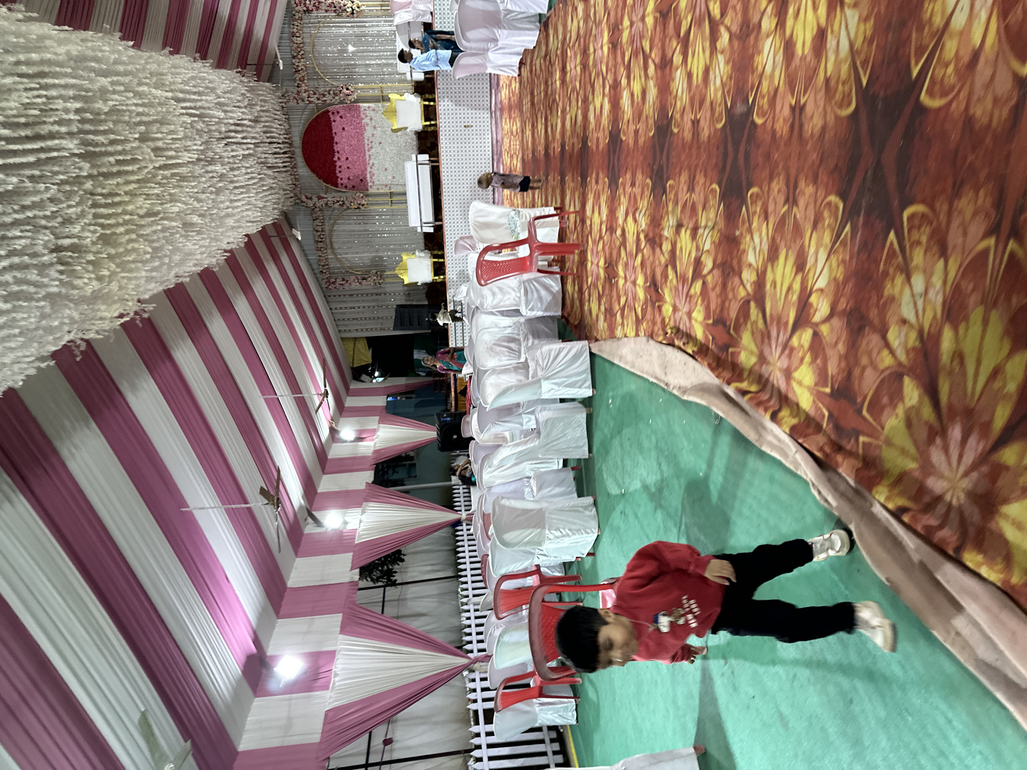 The image shows the interior of a tent in what appears to be Shahapur, India, possibly set up for a wedding or a large social event. The tent's ceiling is decorated with pink and white striped fabric draping and large white fluffy decorative elements. In the background, there is a decorated stage or backdrop featuring floral arrangements and what appears to be red and white circular fabric panels.
Rows of white chairs are set up facing the stage, with some red plastic chairs interspersed among them. A patterned rug covers the floor, transitioning from the tent's green ground covering. Several people are seated in the chairs, and a child wearing a red sweatshirt and dark pants is walking on the green ground covering.