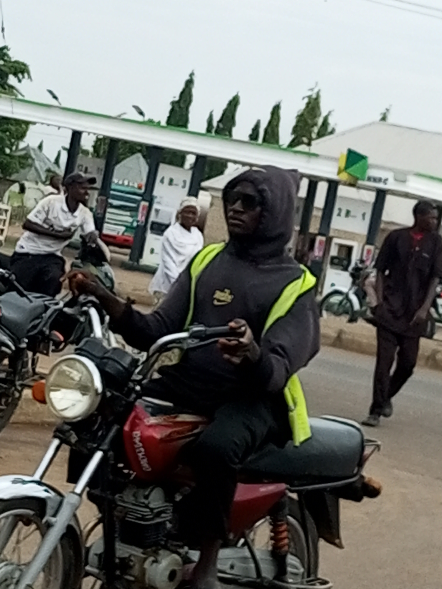 In Gombe, Nigeria, a person wearing a dark hooded jacket with a bright yellow vest over it is riding a motorcycle. The motorcycle has a red fuel tank with white lettering on it, and a chrome headlight. The rider is wearing sunglasses. In the background, there are several gas pumps with a white roof structure above them. A green and yellow diamond-shaped sign is visible on the roof structure. Other people are standing and walking in the background near the gas pumps, some of whom are wearing traditional clothing. The scene appears to be at a gas station on a street.