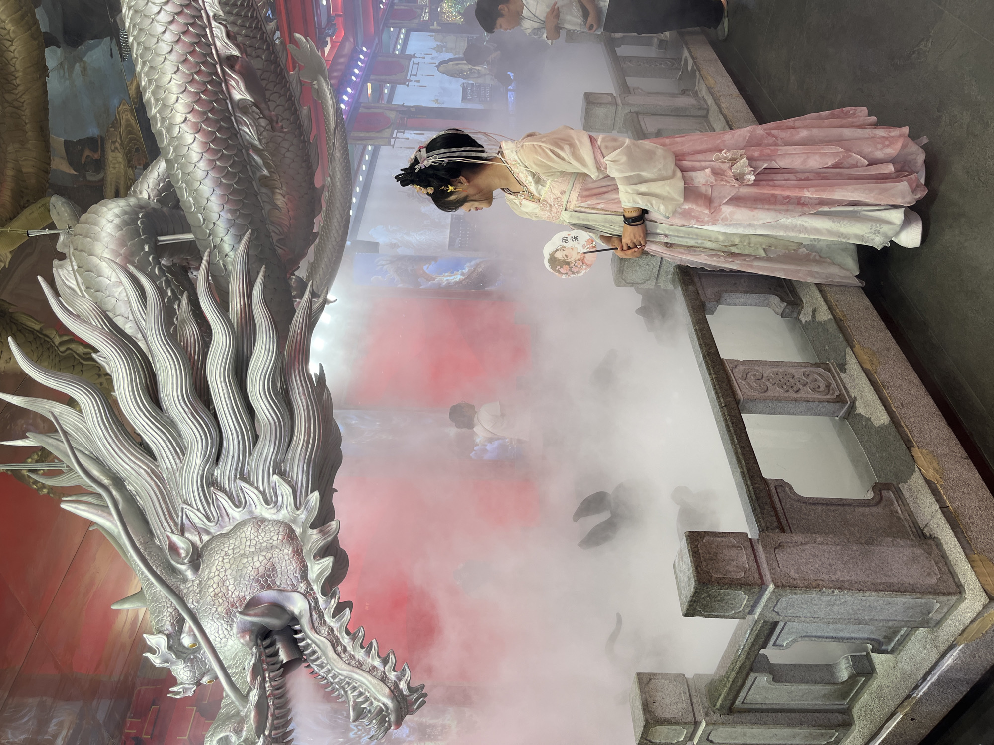The image shows a young woman in traditional Chinese attire standing next to a large, silver dragon statue. The woman is wearing a pink and white dress with intricate details and has her hair styled in an updo with ornaments. She is holding a fan adorned with what appears to be a portrait. The dragon statue is highly detailed, with scales and flowing mane, and seems to be spitting out smoke or fog, which also fills the surrounding area, creating a dramatic and ethereal atmosphere.
The setting appears to be an outdoor or semi-outdoor venue, possibly a park or a themed attraction, with stone structures and traditional Chinese architectural elements visible in the background. There are other people in the background, some observing the scene, others blurred by the fog. The lighting suggests it might be evening or a dimly lit indoor space, with artificial lights illuminating the scene from above. The overall mood is one of fantasy and cultural immersion, with the vibrant colors of the woman's dress contrasting with the metallic sheen of the dragon and the soft haze. There is some text on the fan, but it is not clearly legible.