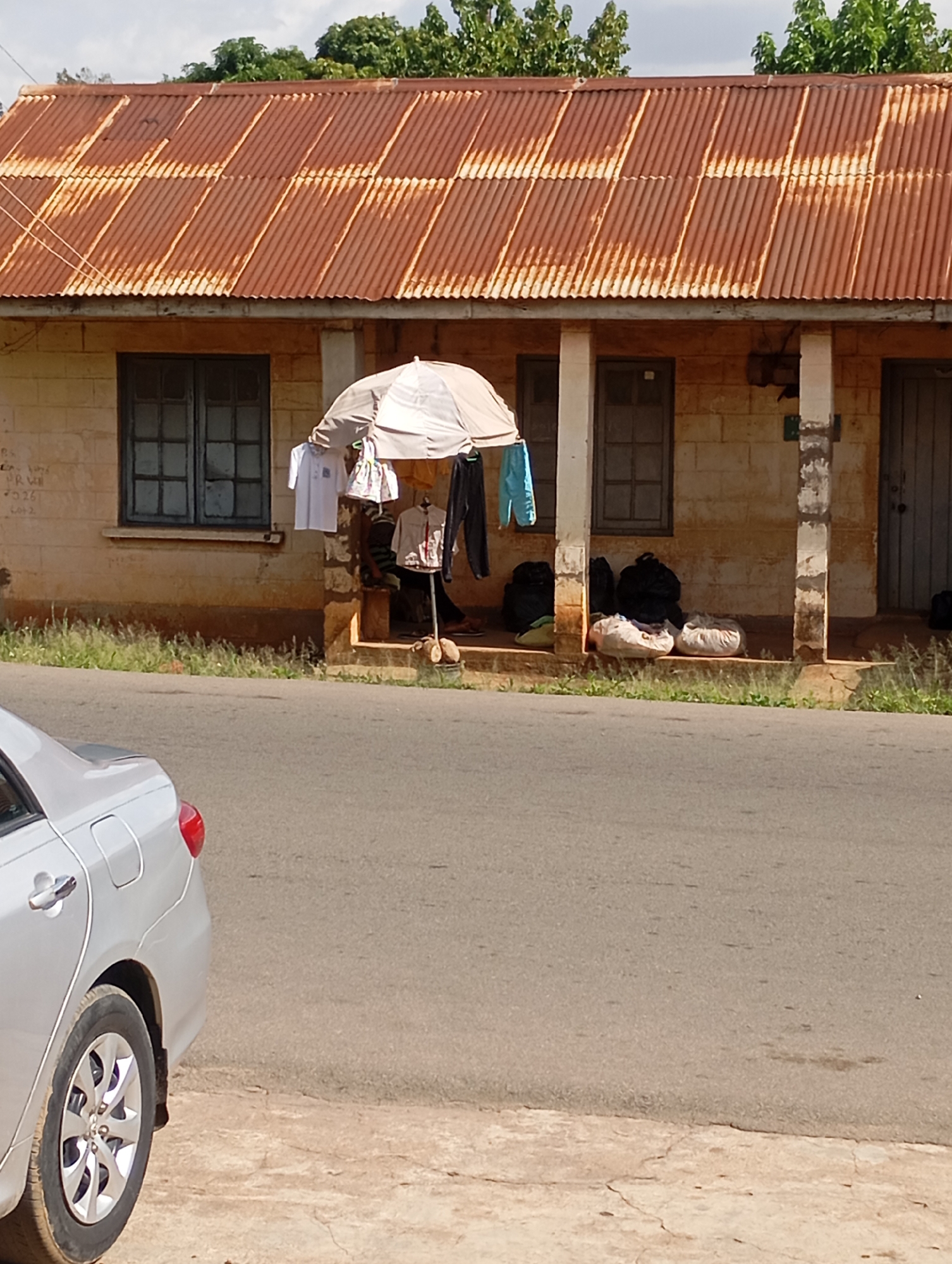 The image shows a roadside scene in Jos, Nigeria. In the foreground, the rear section of a silver car is visible, parked on a paved surface adjacent to a road. The car has a visible wheel and taillight. Behind the car, a building with a rusty corrugated iron roof is present. In front of the building, a stall is set up with clothes hanging on a rack and an umbrella providing shade. Several bags are piled on the ground near the stall. The building itself appears to be constructed from light-colored bricks. Green vegetation, likely grass and trees, is visible in the background. The scene depicts a typical urban or semi-urban setting, possibly a commercial area with street vendors.