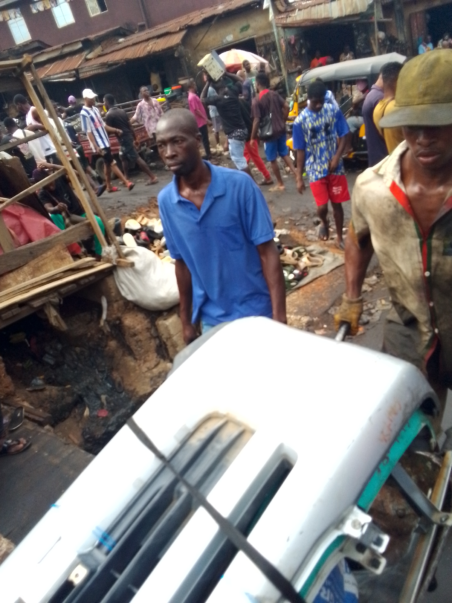 The image depicts a bustling street scene in Onitsha, Nigeria. Two men are pulling a white and teal handcart through a crowded market area. The man in the foreground wears a yellow-brown hat and a dirtied olive green shirt with red trim and work gloves. The other man wears a blue polo shirt. Both men appear to be focused on their task.
The surroundings include:
* A busy open-air market with pedestrians walking in different directions.
* Makeshift stalls and shops with corrugated metal roofs.
* A drainage ditch in the foreground covered in some areas with makeshift wooden structures and littered with debris.
* A yellow motorized tricycle (keke) partially visible in the background, suggesting this is a form of public transportation.
* Miscellaneous objects like bags, bottles, and discarded items are scattered around the ground.
The scene suggests an urban commercial environment, with evidence of both trade and waste. The atmosphere seems active and somewhat chaotic.