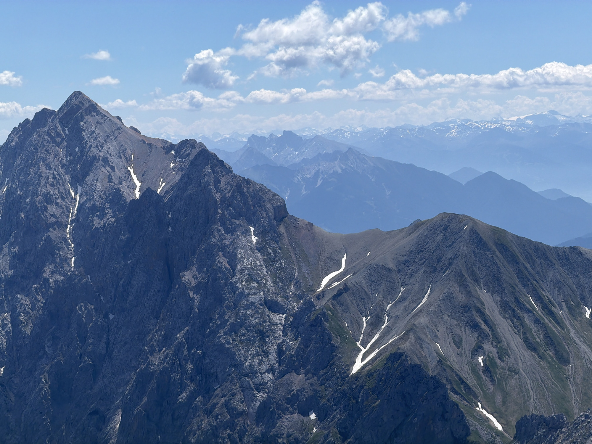 This is a photograph taken from a high vantage point, likely a mountain peak, overlooking a vast mountain range under a clear blue sky with scattered white clouds. The foreground is dominated by a rugged, dark grey mountain peak with sharp, angular features. Patches of white snow are visible in crevices and on the slopes of this foreground mountain.
In the mid-ground and background, successive layers of mountains fade into a hazy blue, indicating significant distance and atmospheric perspective. These distant ranges also show signs of snow on their higher elevations. The overall scene is one of grandeur and natural beauty, typical of alpine landscapes.
Considering the prompt's mention of Garmisch-Partenkirchen, Germany, this photograph is very likely a view from the Zugspitze, Germany's highest peak, or a similar vantage point in the Bavarian Alps. The characteristic jagged peaks, sparse vegetation at higher altitudes, and the layering of distant mountains are consistent with the geography of this region. The lighting suggests it is daytime, possibly late morning or early afternoon, with the sun illuminating the scene from the right. There are no visible people, man-made structures, or specific actions taking place within the frame.